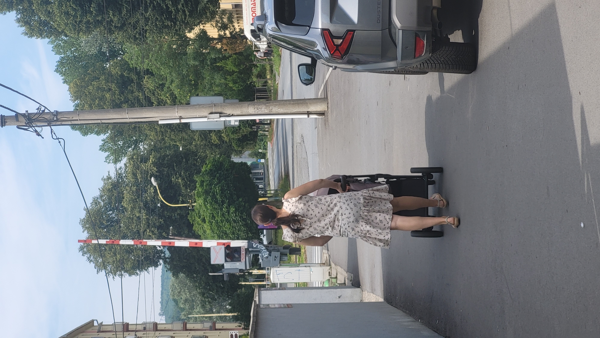 A woman with her back to the camera is pushing a stroller down a sidewalk in Prešov, Slovakia. She is wearing a light-colored, floral-patterned dress and tan sandals. The stroller is dark-colored. To the right of the sidewalk is a grey SUV. Further to the right, across the street, is a white truck with the word "ROMAD" visible on its side. In the background, there are trees, buildings, and a railway crossing gate. The sky is partly cloudy. The scene appears to be a typical urban street on a bright day.