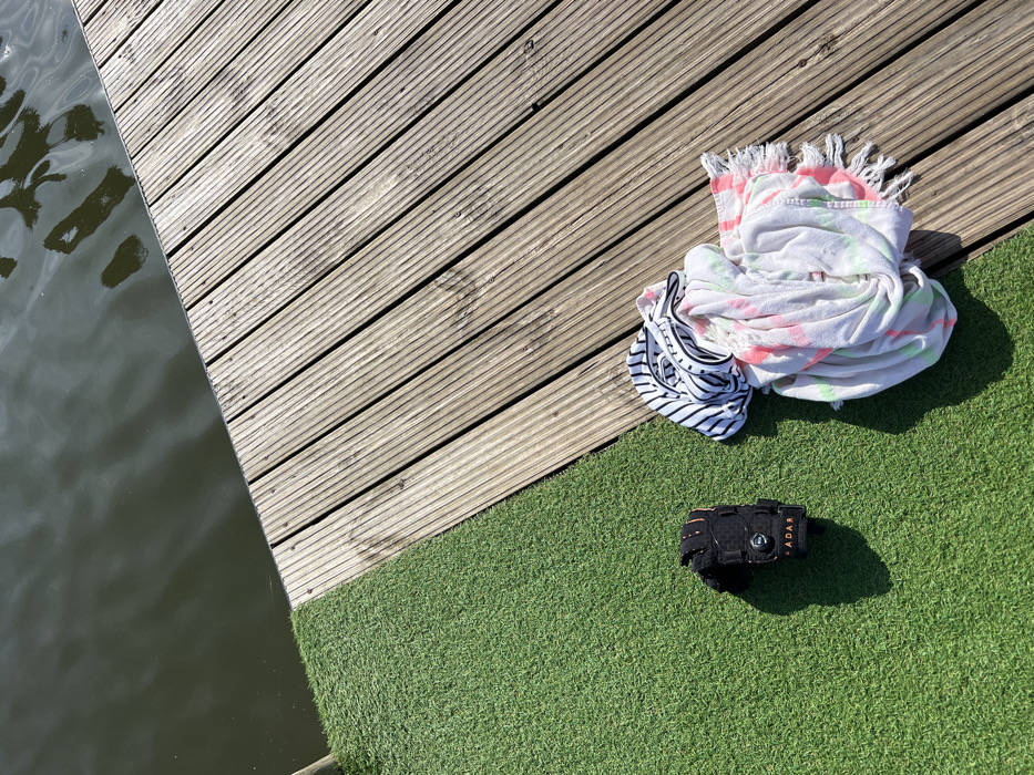 The scene depicts a recreational area adjacent to a body of water in Coruche, Portugal. The image is divided into three distinct zones: a dark, calm body of water in the upper left, a light-colored wooden plank deck running diagonally from the upper right to the lower center, and an area of bright green artificial turf in the lower left. On the artificial turf, a single black watersports glove or wrist support, branded "RADAR" with an orange accent and a central circular fastener, is present. Adjacent to it, partially resting on the wooden deck, is a crumpled pile of textiles. This pile consists of a white garment with black horizontal stripes and black lettering on top, and underneath, a white towel or blanket featuring neon pink and green tie-dye patterns and a fringed edge. Strong overhead sunlight is indicated by the distinct shadows cast by the objects onto the artificial turf and the reflections on the water surface. No individuals are visible in the frame.