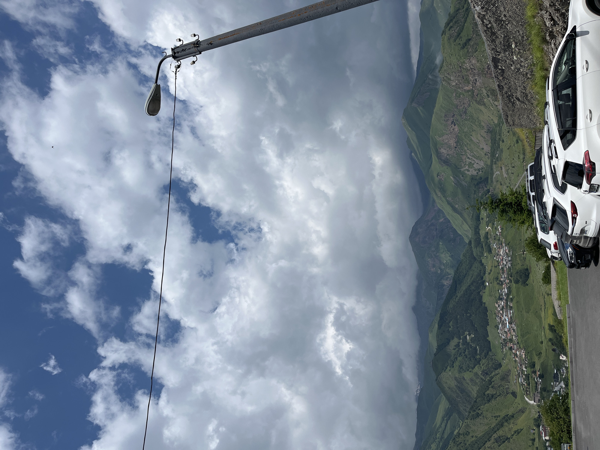 This is a landscape photograph taken in Stepantsminda, Georgia. The image features a panoramic view of green, rolling mountains, a village nestled in the valley, and a cloudy sky. In the foreground, there is a row of parked white SUVs and a black SUV. A utility pole with a street light and electrical insulators stands on the right side of the frame, with a dark electrical wire stretching across the upper portion of the image. The mountains in the background are covered in lush green vegetation, with some rocky outcrops visible. The village in the valley consists of numerous houses with red rooftops, spread across the green slopes. The sky is filled with a mix of white and dark gray clouds, with patches of blue sky visible. The overall impression is one of a serene mountain town on a partly cloudy day.