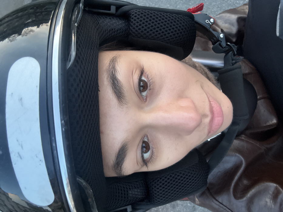 A close-up portrait depicts an individual, appearing female, situated outdoors in the city of Paris, France. The subject wears a glossy black open-face motorcycle helmet with a chrome trim and a white rectangular sticker affixed to the upper front. The helmet's black mesh internal padding and ear covers are visible. The individual's face, characterized by a medium skin tone, dark brown eyes, and prominent eyebrows, is positioned centrally, looking directly into the camera. The lips are slightly parted. Below the helmet's chin strap, a brown leather jacket is visible, underneath which a lighter, possibly beige, garment is present. Reflections on the helmet surface suggest an urban environment with blurred architectural elements and a clear sky.