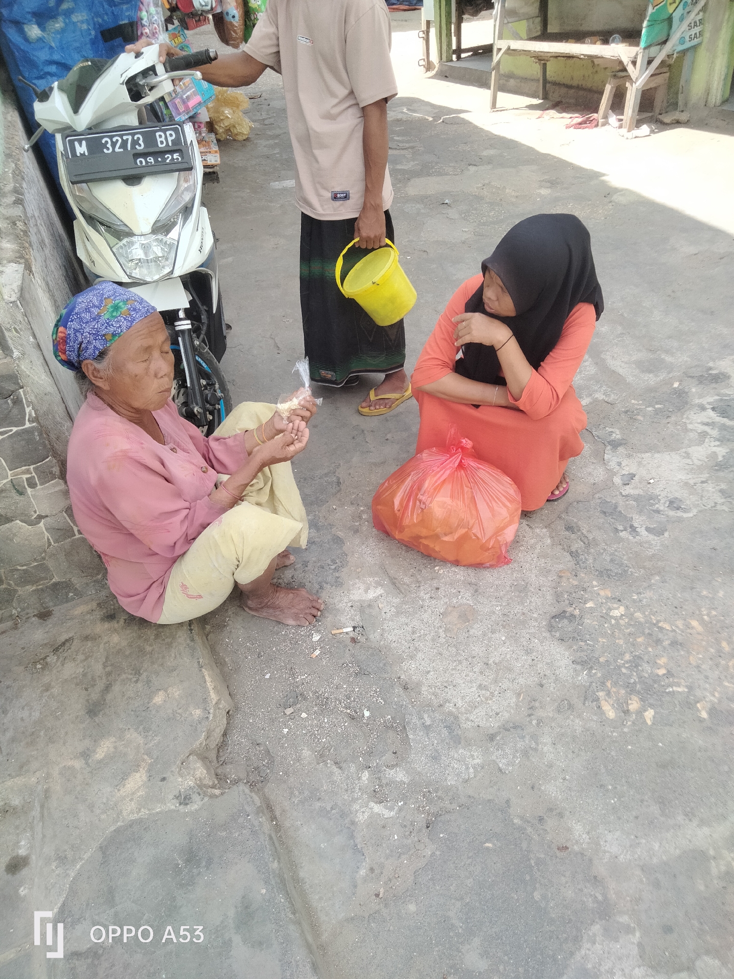In a street scene in Badung, Indonesia, an elderly woman wearing a pink shirt and a blue headscarf sits on the ground next to a white motorcycle, holding a small plastic bag of something to eat. Beside her, a woman in an orange dress and a black hijab kneels on the pavement, her arm resting on her knee. In the background, a man in a light brown t-shirt and a black sarong stands holding a yellow bucket. The ground is paved with concrete, and sunlight casts shadows across the scene.