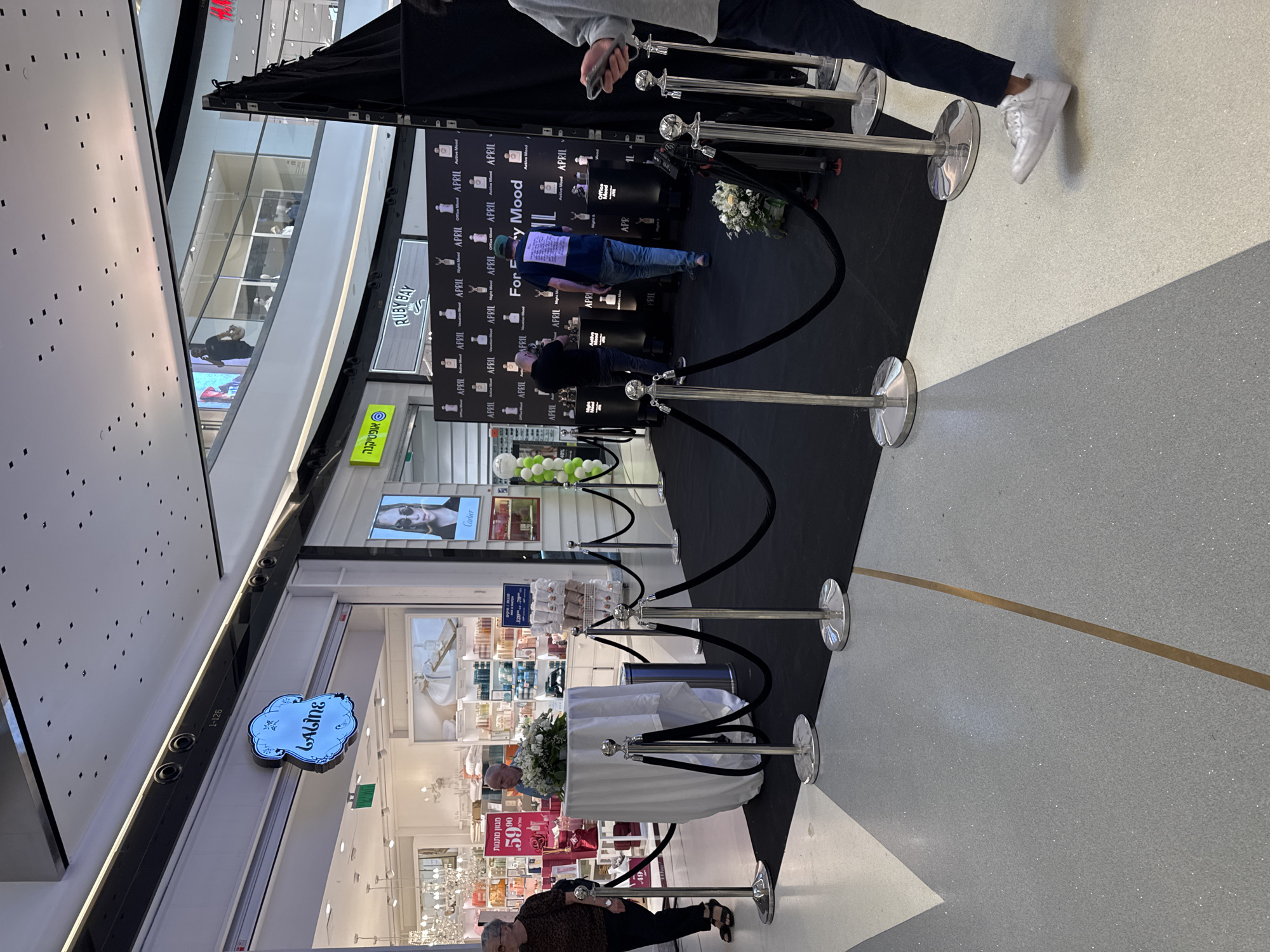In Tel Aviv, Israel, a retail establishment featuring a "Laline" sign is visible, with a display setup in the foreground. This display consists of a black carpeted area cordoned off by chrome stanchions and black velvet ropes. The display itself is a black backdrop featuring multiple perfume bottles arranged in rows, each with the text "APRIL" and "For Every Mood" indicating different scent profiles. There are three men interacting within the display area. One man, wearing a blue t-shirt and a green cap, is facing away from the camera and appears to be looking at the backdrop. Another man in a black shirt is also facing away, looking at something to his left. The third man, partially visible on the right, is wearing a gray long-sleeved shirt and dark pants, and is holding a mobile phone. A white tablecloth-covered display table with a floral arrangement is situated to the left of the main display, with a woman in a dark patterned shirt and sandals standing to its left. The floor of the shopping mall is a speckled gray and white terrazzo pattern.