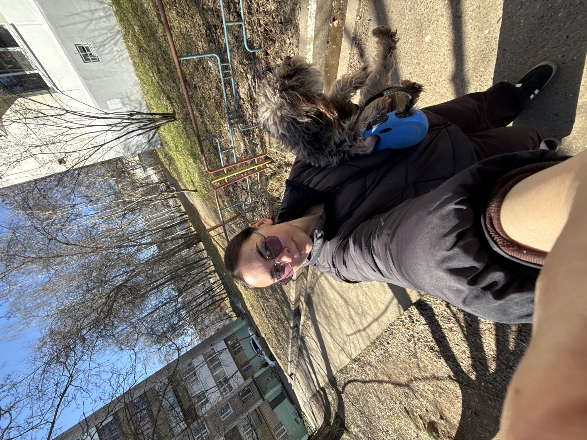 A person is holding a dog on a leash, on a sidewalk in an urban setting. The person is wearing sunglasses and a dark jacket, while the dog appears to be a medium-sized, fluffy breed. The location is identifiable as Chaikovsky, Russia, based on the visible Soviet-era apartment buildings in the background. Additional elements in the scene include a playground apparatus to the left of the dog and sidewalk under the feet. The weather appears to be sunny.