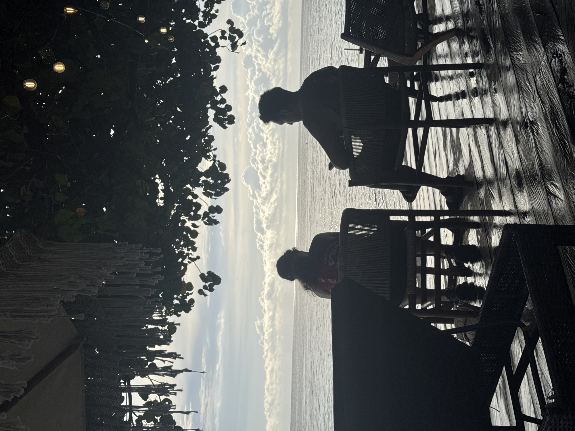 The image depicts two individuals seated on chairs, possibly made of wicker, on what appears to be a wooden deck. They are silhouetted against a bright sky and ocean view, suggestive of a sunset or sunrise. A tree with string lights is visible on the left side of the frame, along with a partially visible macrame canopy or decoration. The scene likely represents a coastal or beachside setting in Nusa Penida, Indonesia.