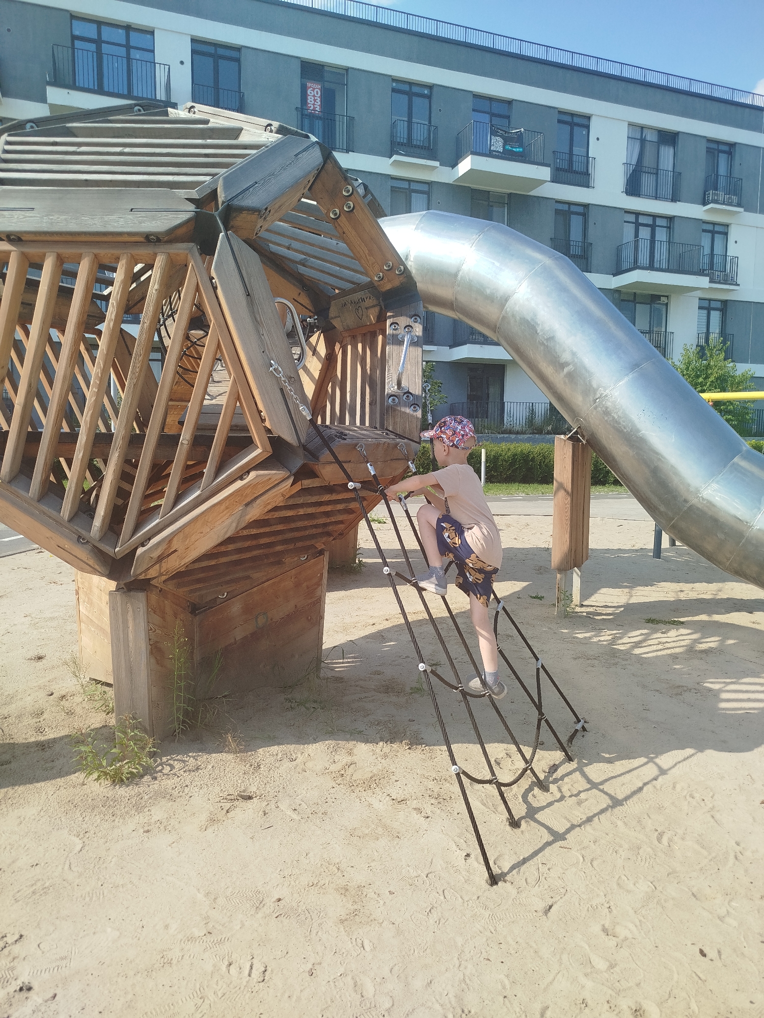The image shows a boy climbing a rope ladder on a playground in Tyumen, Russia. The playground equipment is made of wood and metal, and includes a large slide. The boy is wearing a t-shirt, shorts, and a hat. He is climbing the rope ladder with his right hand and left foot. In the background, there is a modern apartment building. The weather is clear and sunny.