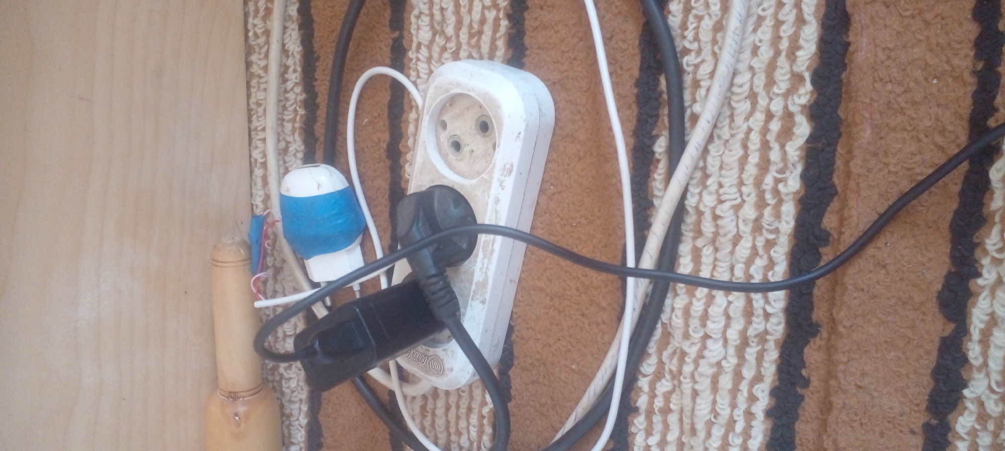 The image shows a multi-plug electrical outlet in what appears to be a domestic setting. The outlet is white and attached to a wooden wall with patterned rug next to it. It has a black plug inserted into one of its sockets. Additional plugs and wires are clustered around the outlet. One plug is white with blue electrical tape. There is a wooden object with what looks like jewelry next to the taped plug.
The location in Parkent, Uzbekistan is not discernable from the image.