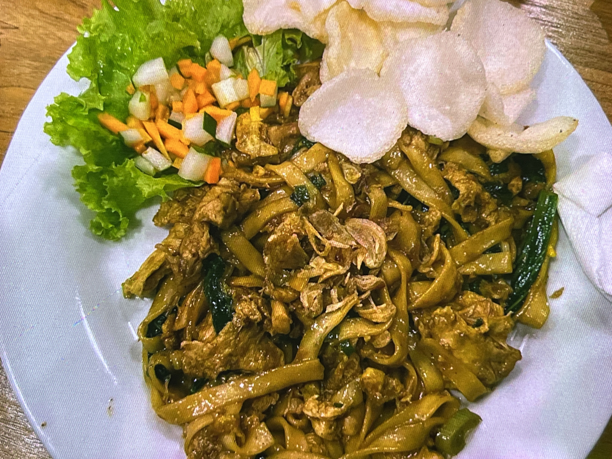 A close-up shot shows a plate of Indonesian food, likely Mie Goreng, served on a white textured plate, placed on a wooden table. The dish features wide, flat noodles coated in a dark, savory sauce, interspersed with pieces of meat and dark green vegetables. Garnishes include a crisp lettuce leaf topped with a diced salad of carrots and cucumbers, and several translucent white crackers (krupuk) arranged on the side. The lighting is warm and direct, creating highlights on the glossy noodles and suggesting an indoor setting, possibly a restaurant or home. The image is framed from a top-down perspective, focusing entirely on the food presentation.