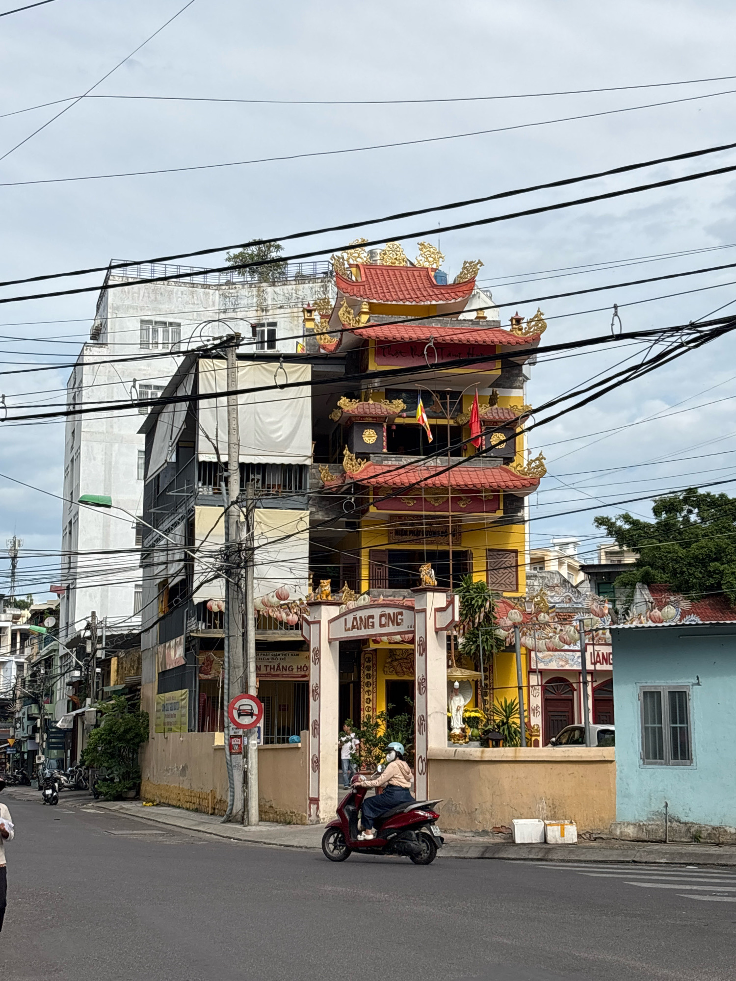 In a street scene in Nha Trang, Vietnam, a tall, narrow, yellow and red temple structure stands out amidst other buildings. The temple features tiered, ornate roofs with gold dragon decorations and red tiles. A large archway with the inscription "LANG ONG" (meaning "Old Man Temple") leads into the temple. On the right side of the temple, a light blue single-story building with a window is visible.
In front of the temple, a person rides a red scooter, wearing a light brown jacket, blue skirt, and a white helmet. Several parked scooters and motorcycles are seen further down the street to the left. A traffic sign indicating no entry for cars is present on a pole. The street is paved, with a pedestrian crossing marked. Utility wires crisscross the sky above the buildings. The sky is overcast with some clouds.