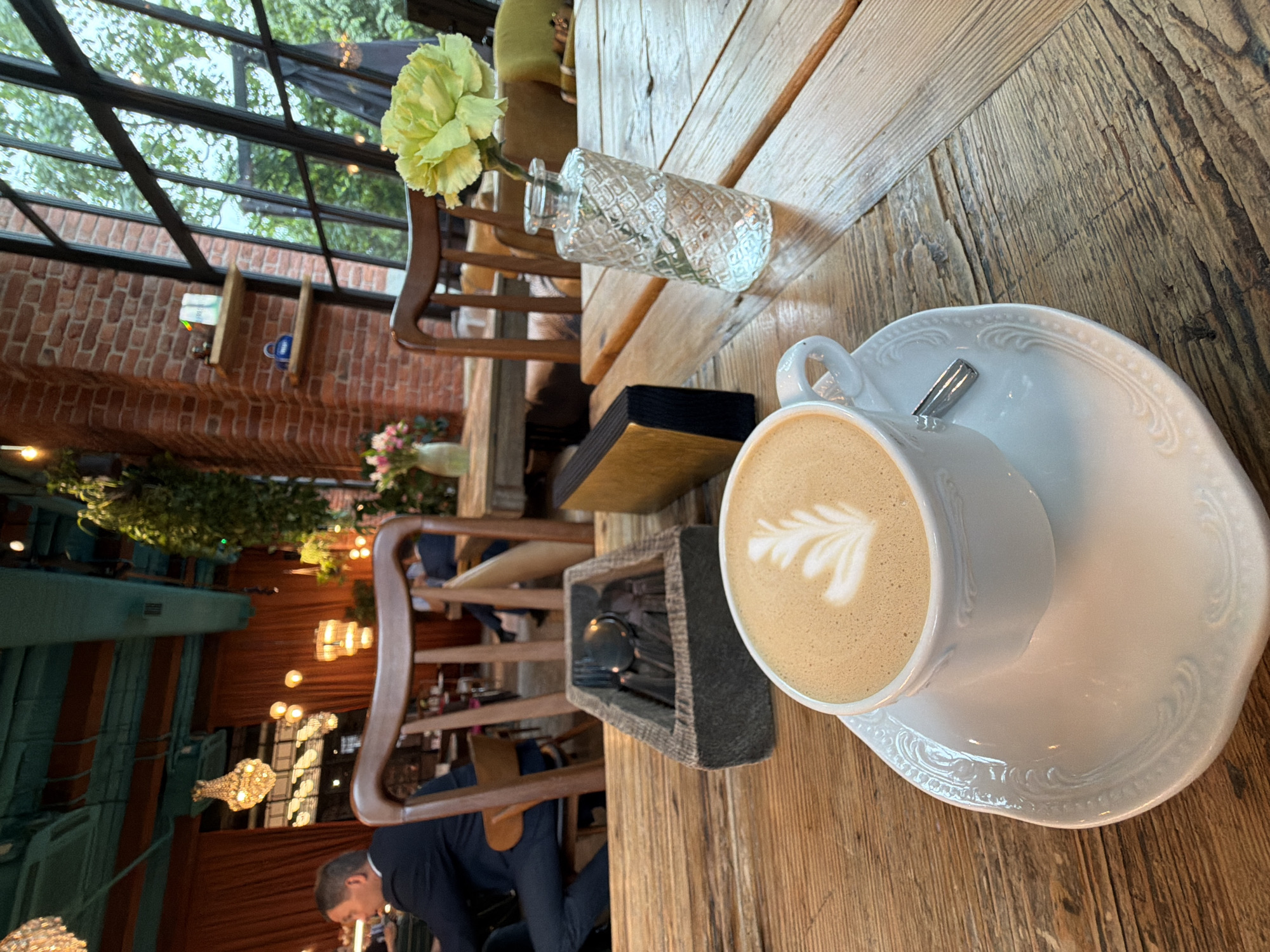 The image depicts a cozy cafe interior. In the foreground, a white cup of coffee with latte art rests on a saucer on a wooden table. Beside the cup is a grey wooden holder containing cutlery. To the right of the coffee, a small glass vase holds a single yellow flower. The background reveals a seating area with wooden chairs and tables, and large windows offering a view of greenery outside. The cafe has exposed brick walls and industrial-style ceiling elements. The overall ambiance is warm and inviting. This scene could be located in Saint Petersburg, Russia, as such cafes are common in the city, known for its blend of historical architecture and modern establishments.