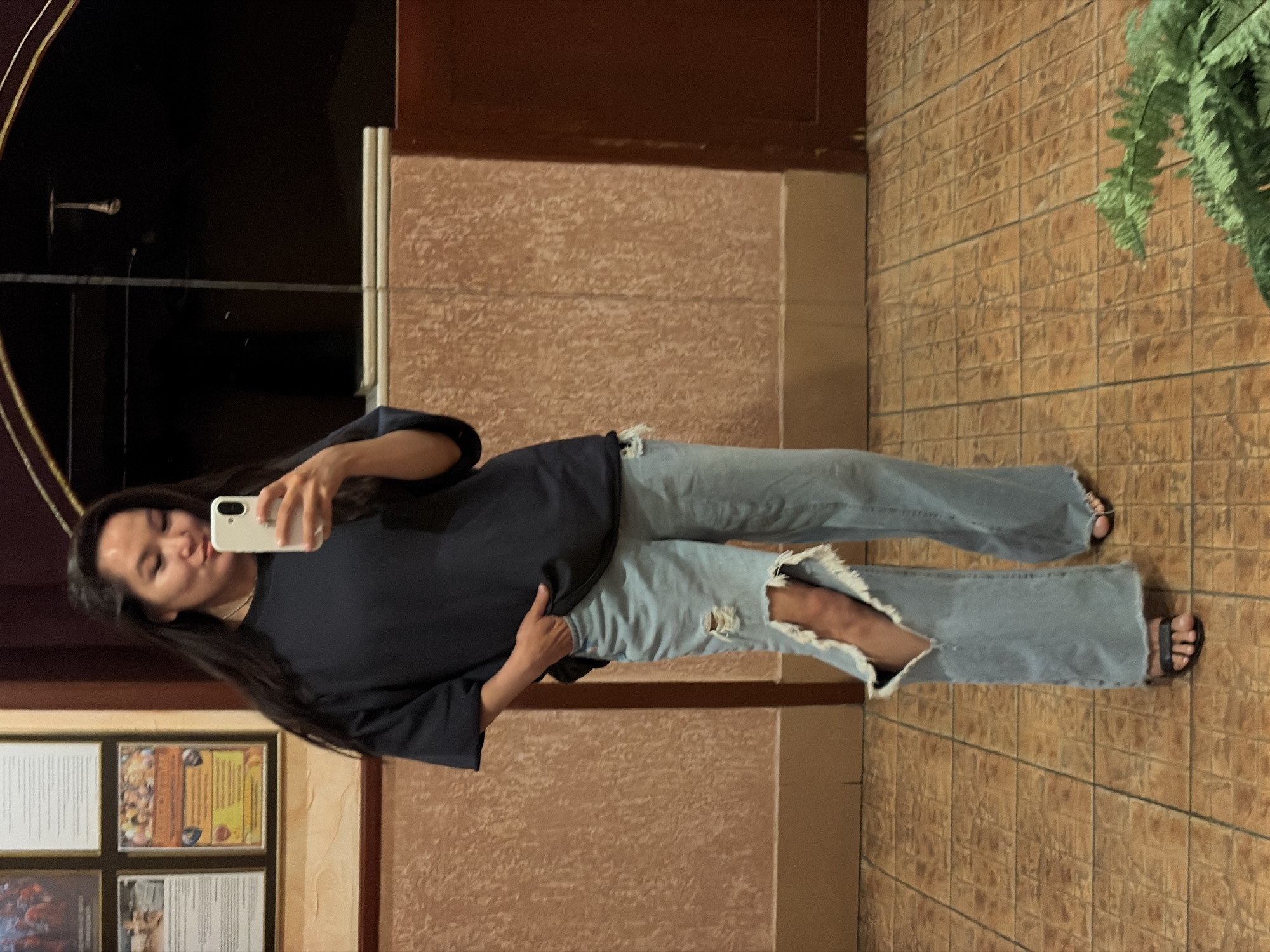 A woman is standing indoors, posing for a selfie. She's wearing a dark blue, oversized T-shirt and ripped blue jeans. She is also wearing black sandals. She is holding a white smartphone up to take the picture. The background features a wall with brown textured tiles, a wooden door frame, some framed posters, and a decorative green plant. The location is likely Chaikovsky, Russia.