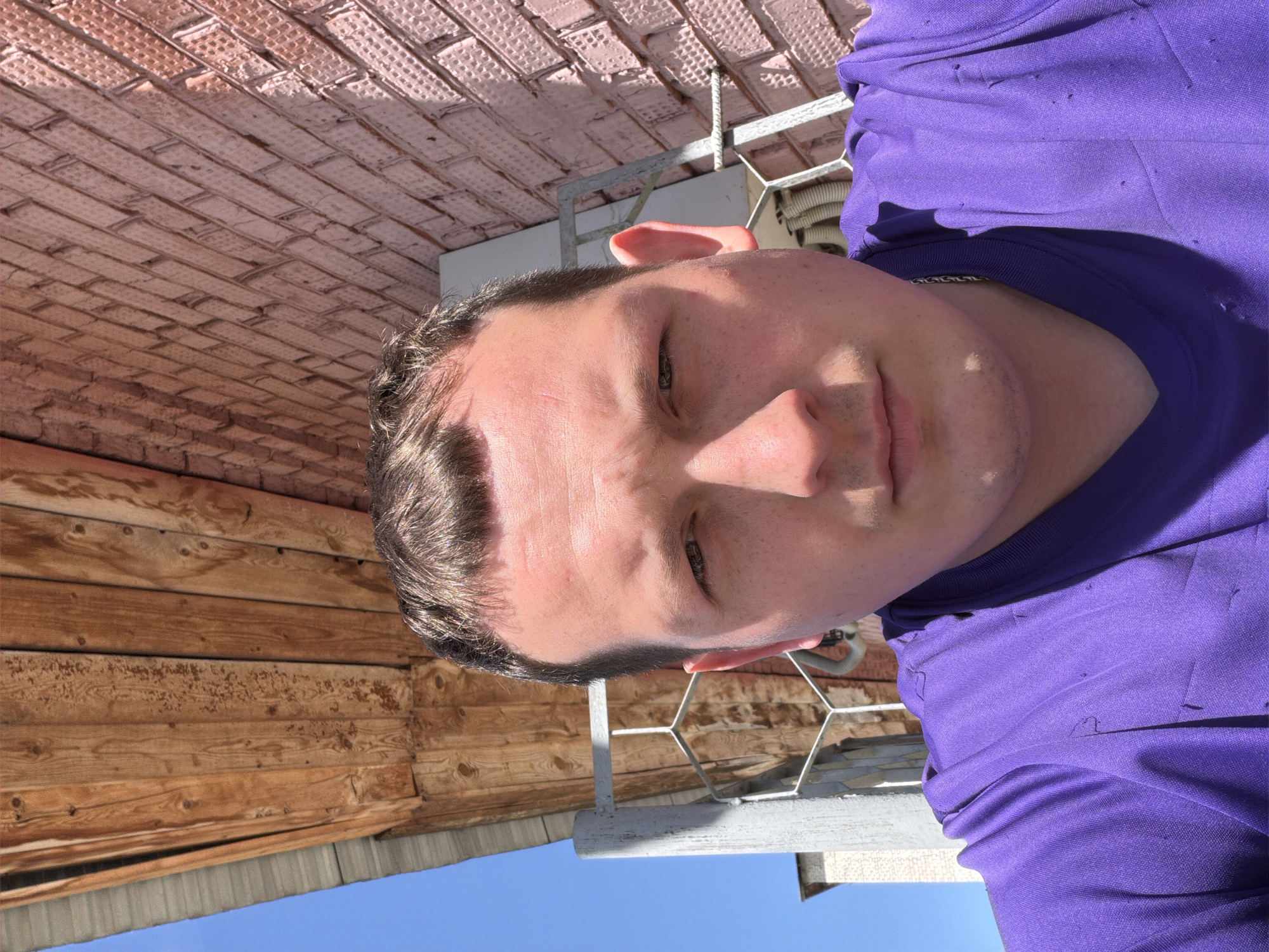 A close-up, upside-down photograph shows a person wearing a purple shirt. The background includes a wooden structure, a brick ceiling, a metallic railing, and a blue sky. The location is unknown and cannot be determined as Chaikovsky, Russia.