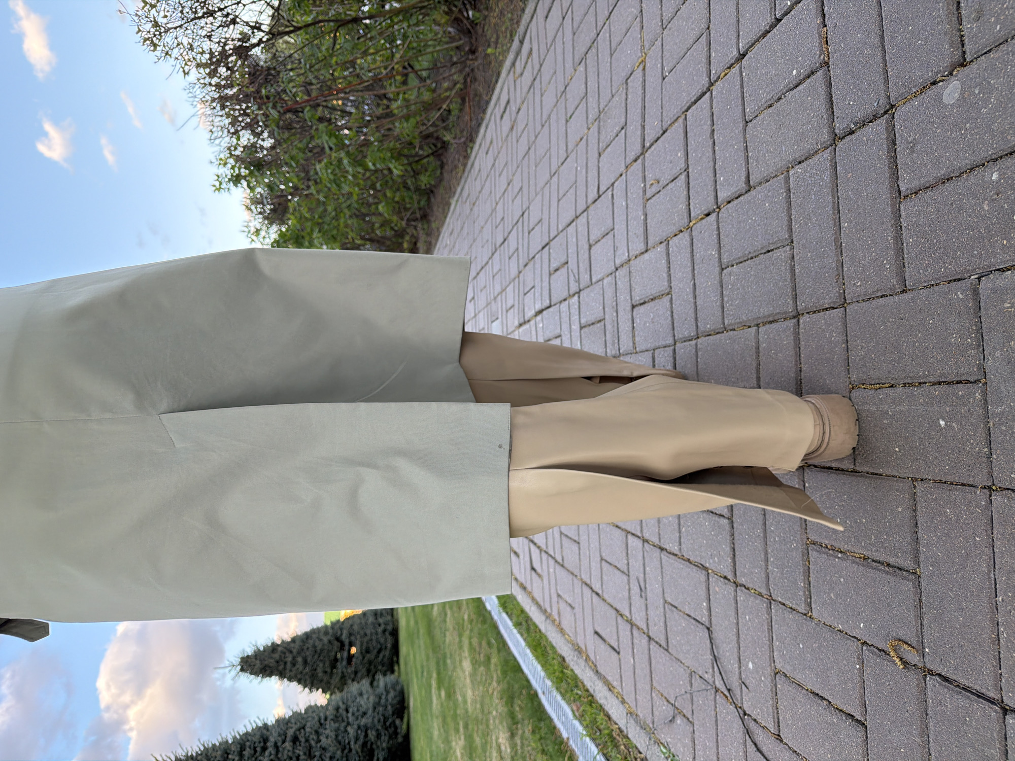 A person stands on a brick walkway in Izhevsk, Russia. They are wearing tan boots and a light green trench coat. The scene is outdoors with trees and vegetation visible in the background. The sky is blue with some clouds.