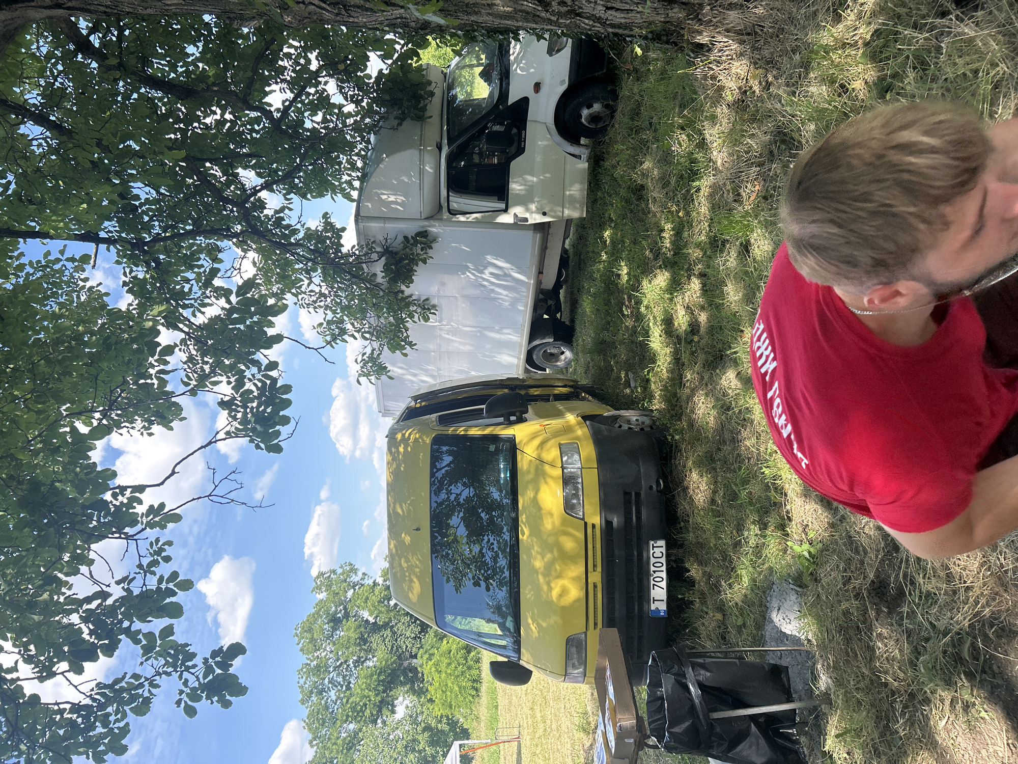 The image depicts an outdoor scene, likely in Lovets, Bulgaria, on a sunny day with scattered clouds. Two vans are parked on a grassy area, partially shaded by a large tree. In the foreground, the back of a person wearing a red t-shirt is visible. The t-shirt has white text on the back, though it is difficult to read fully. A silver chain is visible around the person's neck.
The first van is yellow and faces forward, with its headlights and windshield clearly visible. The license plate reads "T 7010 CT". Next to it, a larger white van is parked, facing the same direction, with its cargo area extending behind the cab. The scene appears to be in a rural or semi-rural setting, with trees and greenery surrounding the vehicles. The sun casts shadows, suggesting it is likely midday. The overall atmosphere seems relaxed and casual, with the person in the red shirt appearing to be part of the scene, perhaps taking a break or observing the surroundings. The presence of a black trash bag on a stand near the yellow van might indicate some form of gathering or activity.