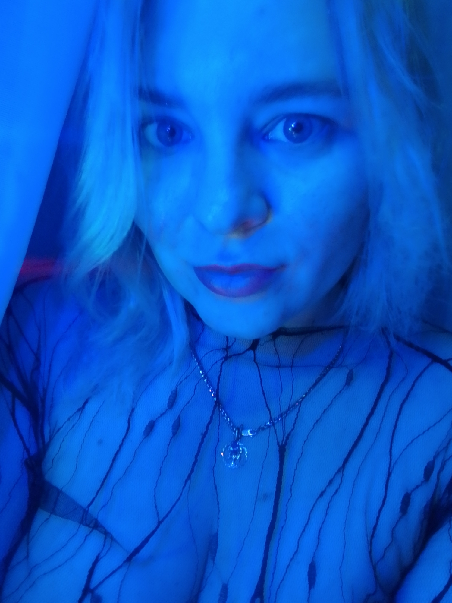 A close-up of a person's face and upper torso is illuminated by blue light. The person has fair skin, light hair, and blue eyes. They are wearing dark lipstick and a delicate silver necklace with a circular pendant. The individual is wearing a sheer black top with a geometric pattern. The background is indistinct due to the heavy blue lighting. There is no indication of the location of the city of Izhevsk, Russia.