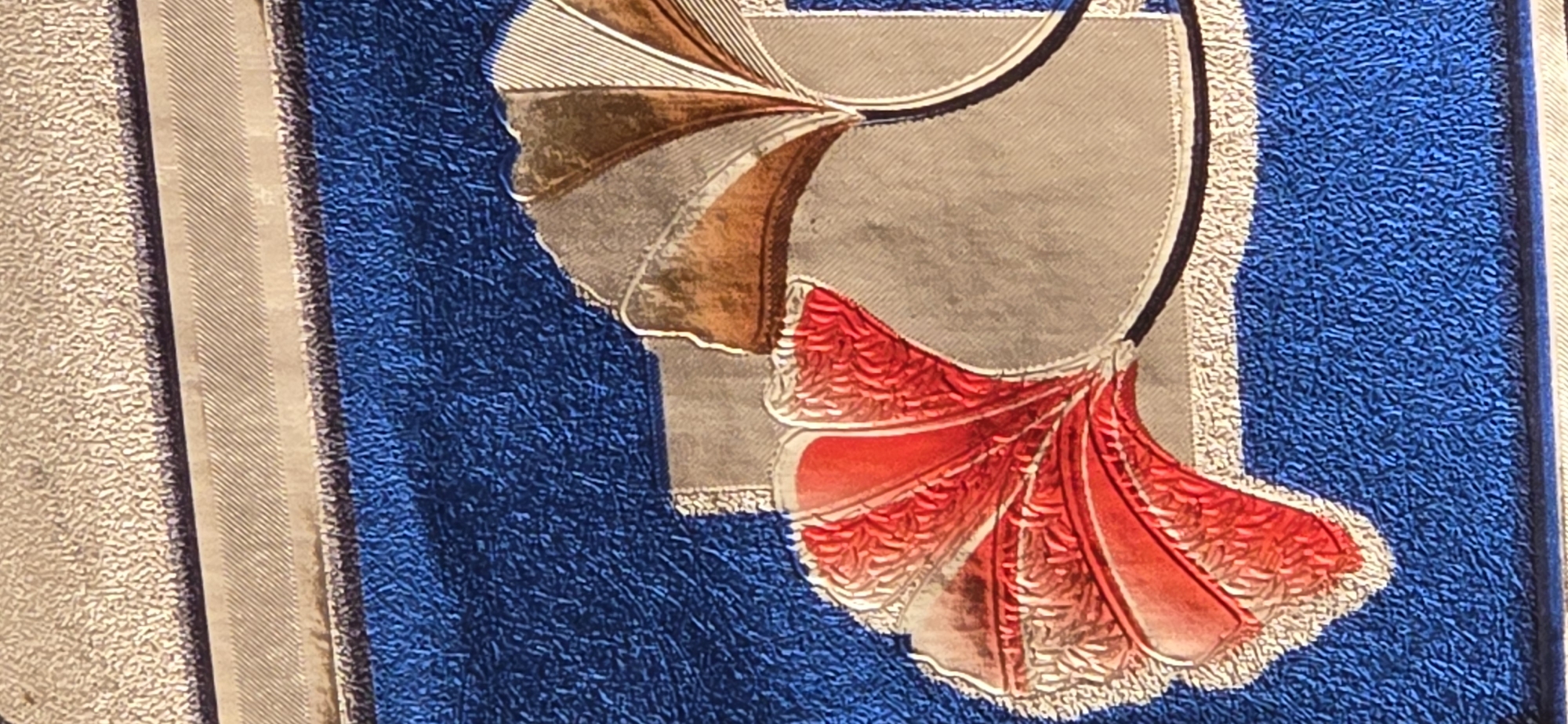 The image shows a close-up detail of a textile or carpet with a textured surface. The design features stylized, fan-shaped elements in red and gold, with a blue background that has a rough, tufted texture. There are also sections with a lighter, possibly beige or off-white, textured material and a thin, ridged band. The arrangement suggests an ornamental or decorative pattern, possibly from a rug, tapestry, or embroidered piece. The lighting creates shadows that emphasize the raised texture of the design. The specific location in Tel, Azerbaijan is not determinable from the visual information provided.