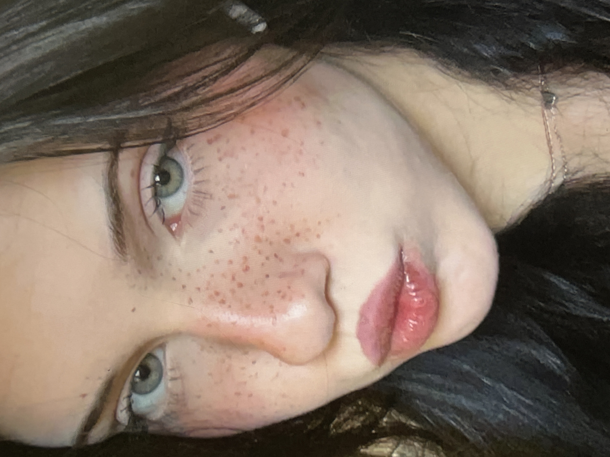 This close-up, side-profile media file features a person's face, oriented sideways. The subject has fair skin with numerous light brown freckles scattered across their cheeks and nose. Their eyes are a striking blue-green color, with visible eyelashes and subtle makeup. The person's lips are full and painted with a glossy, pinkish-red lipstick, creating a wet appearance. Dark, long hair frames the face, with strands visible in the foreground. A delicate silver necklace with a small, dark pendant is visible around the person's neck, resting on their skin near the jawline. The lighting appears to be soft, casting subtle shadows and highlighting the texture of the skin and the gloss on the lips. The overall mood is intimate and artistic, with the focus solely on the facial features. No text is visible in the image. The location context of Cupra Marittima, Italy, suggests a possible connection to this region, though no direct visual cues confirm this.