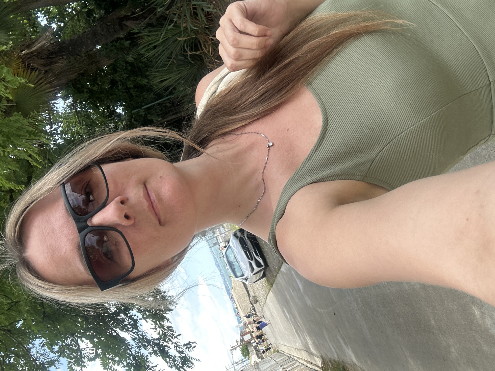 In Sochi, Russia, a woman with blonde hair and sunglasses takes a selfie outdoors. She is wearing a green tank top and a silver necklace with a small pendant. In the background, a silver car is parked on a gravel area next to a paved walkway. Several people are visible in the distance, some walking and others with bicycles. Lush green trees and foliage surround the scene, with a body of water and a distant coastline under a cloudy sky.