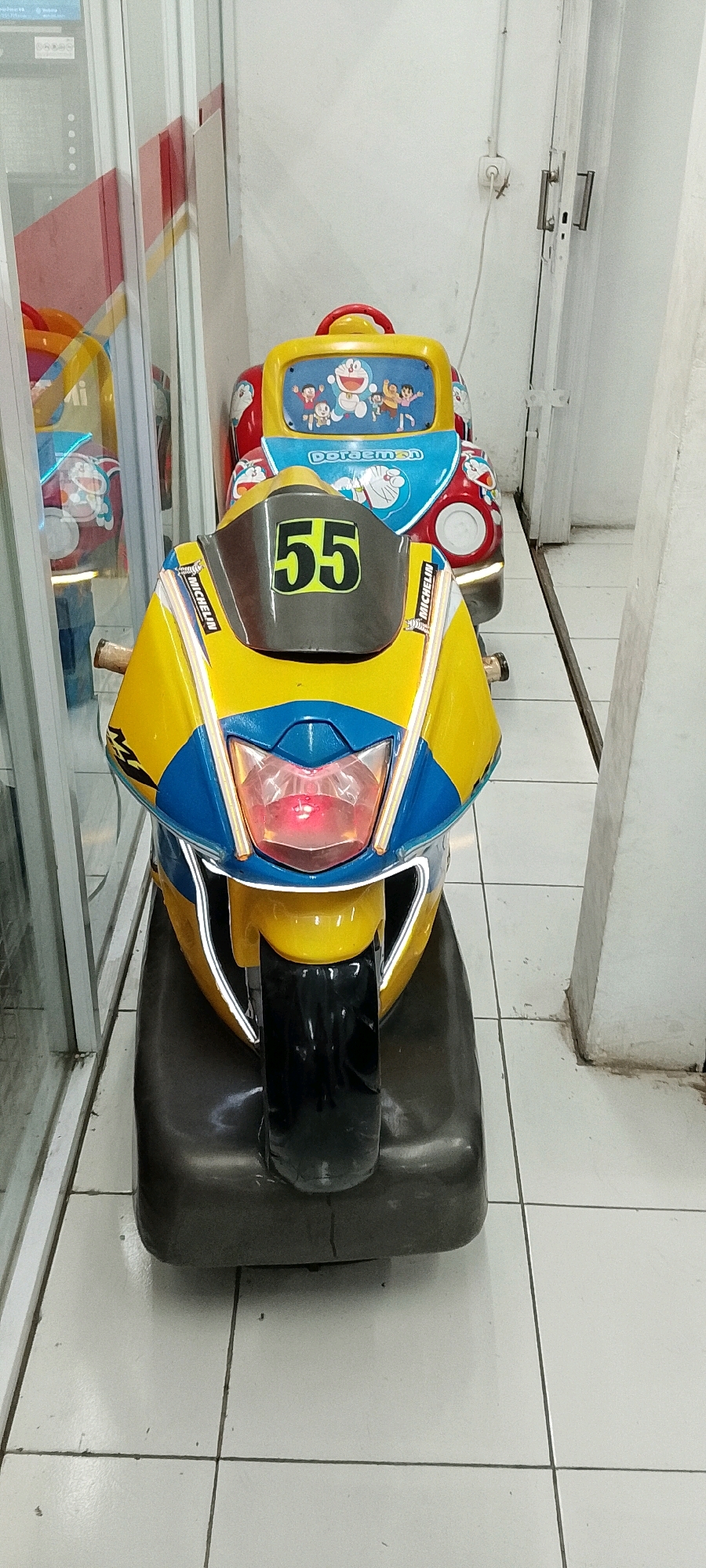 The image shows two children's amusement ride-on toys, a yellow and blue motorcycle in the foreground and a yellow and red Doraemon-themed car behind it, positioned in an indoor space with tiled flooring. The motorcycle has the number "55" prominently displayed on its front. The car features characters from the anime series Doraemon. The toys are stationary, and no people are present in the immediate vicinity. The environment appears to be a clean, indoor public space, potentially an arcade or entertainment center. The background shows a white wall and a door. The overall impression is one of a well-lit, accessible area designed for children's entertainment.