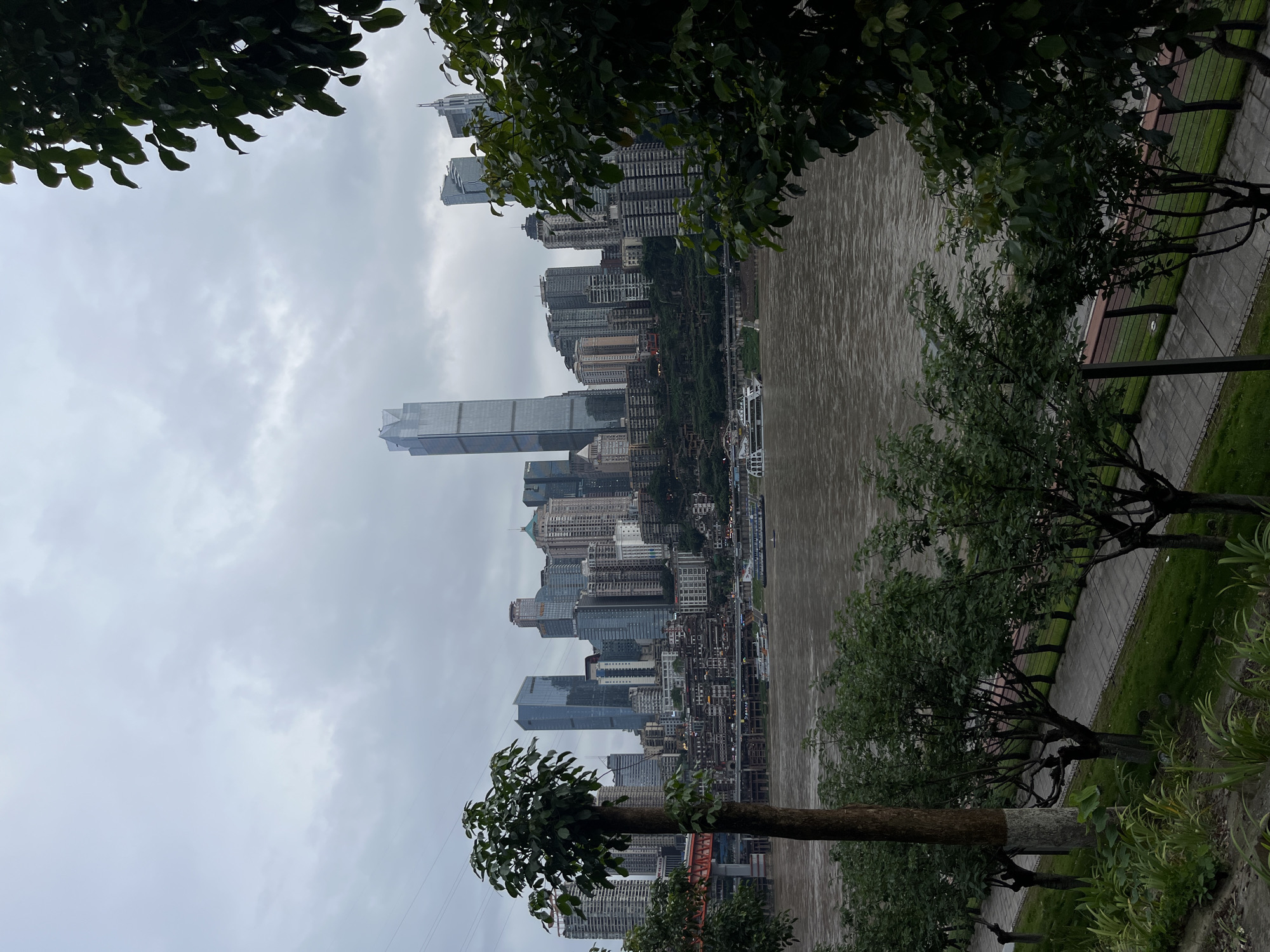 The image shows a cityscape on a cloudy day, partially obscured by trees in the foreground. A wide, muddy river separates the viewer from the city. The city consists of numerous tall buildings, including modern skyscrapers. The area is 江北城街道, China. A paved walkway is visible along the riverbank on the viewer's side, with grass and trees separating it from the river.