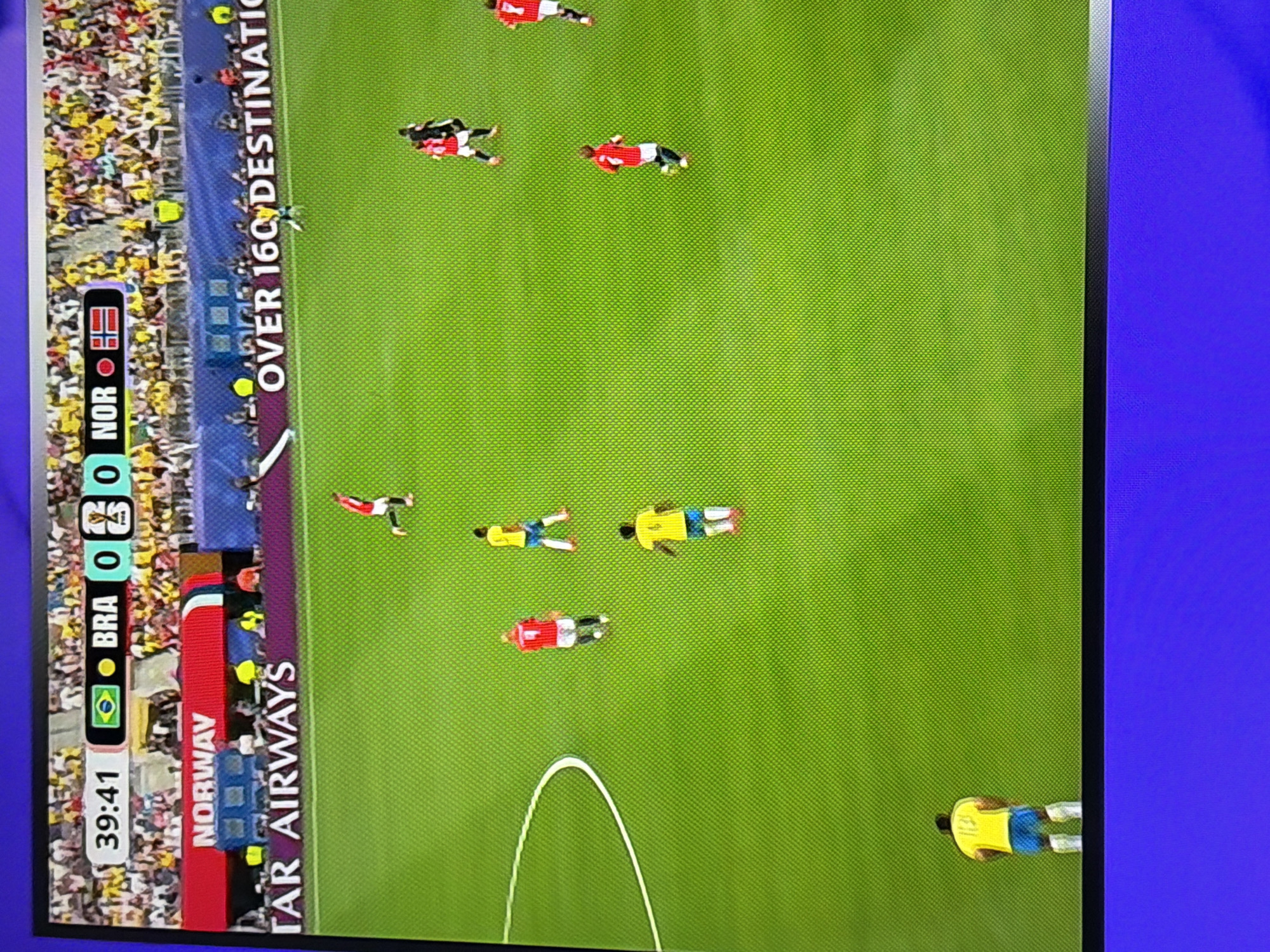 The image shows a soccer match being broadcast on a television screen. The score displayed is Brazil 0, Norway 0 at 39:41. The field is green, and several players in red and yellow uniforms are visible. In the background, spectators are in the stands, and advertising banners for "Norway" and "Qatar Airways" are visible. The television screen is placed on a purple surface. The scene is a still image captured from a television broadcast, not an actual location in Ognianovo, Bulgaria.