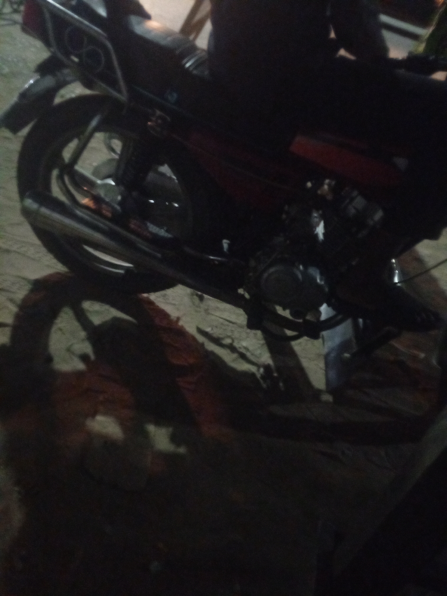 The image shows the rear portion of a motorcycle, likely parked on an uneven surface, potentially dirt or gravel, in Mbribit, Nigeria. The motorcycle is dark in color, with red accents visible on the body panel. The exhaust pipe is prominently displayed. Part of the engine is visible. The image appears to be taken at night or in low light conditions, evidenced by the strong shadows and lack of detail in the background. A person is partially visible on the motorcycle.