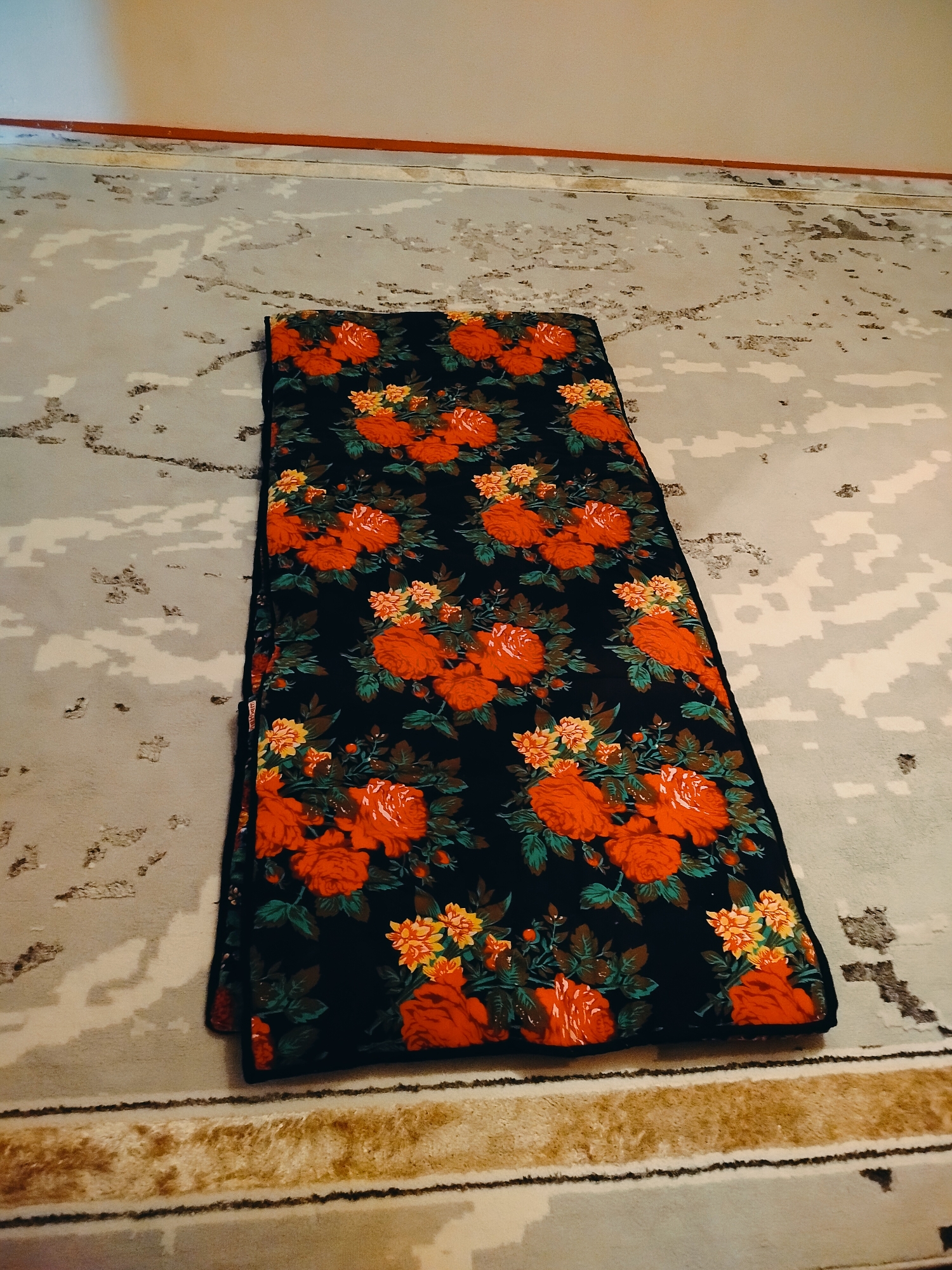 A floral-patterned blanket, predominantly black with red and orange roses and yellow accents, is laid out on a carpet. The carpet has a mottled grey and white pattern, with a striped border visible at the bottom. The blanket is folded lengthwise. The environment appears to be indoors, possibly a room in a residential building in the city of Көлкент, Kazakhstan, as suggested by the wall and baseboard details. The lighting is dim, casting a slight shadow.