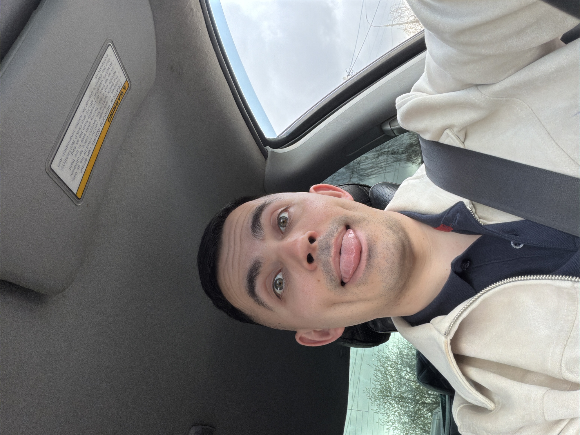 A young man with short dark hair and light eyes is captured in a playful self-portrait, sticking his tongue out and looking directly at the camera. He is seated inside a car, wearing a dark polo shirt beneath a light-colored, possibly off-white, zipped jacket. A dark seatbelt is fastened across his chest and shoulder.
The setting is the interior of a vehicle, possibly in Izhevsk, Russia, viewed from a slightly tilted perspective as the original image is rotated. Above his head, a sunroof is visible, revealing an overcast sky and the faint outlines of bare tree branches and power lines outside. To the left, a car's sun visor is flipped down, displaying a prominent yellow warning label. The car's headliner and parts of the interior are visible, suggesting a functional, rather than luxurious, vehicle.
The visible text on the sun visor reads: "A WARNING DEATH OF SERIOUS INJURY can occur. The BACK SEAT is the SAFEST place for children. Children 12 and under can be killed by the air bag. NEVER put a rear-facing child seat in the front. Sit as far back as possible from the air bag. ALWAYS use SEAT BELTS and CHILD RESTRAINTS. See the Owner's Manual for further information and explanations."
The time of day appears to be daytime under an overcast sky. The man's expression conveys a sense of lightheartedness and humor.