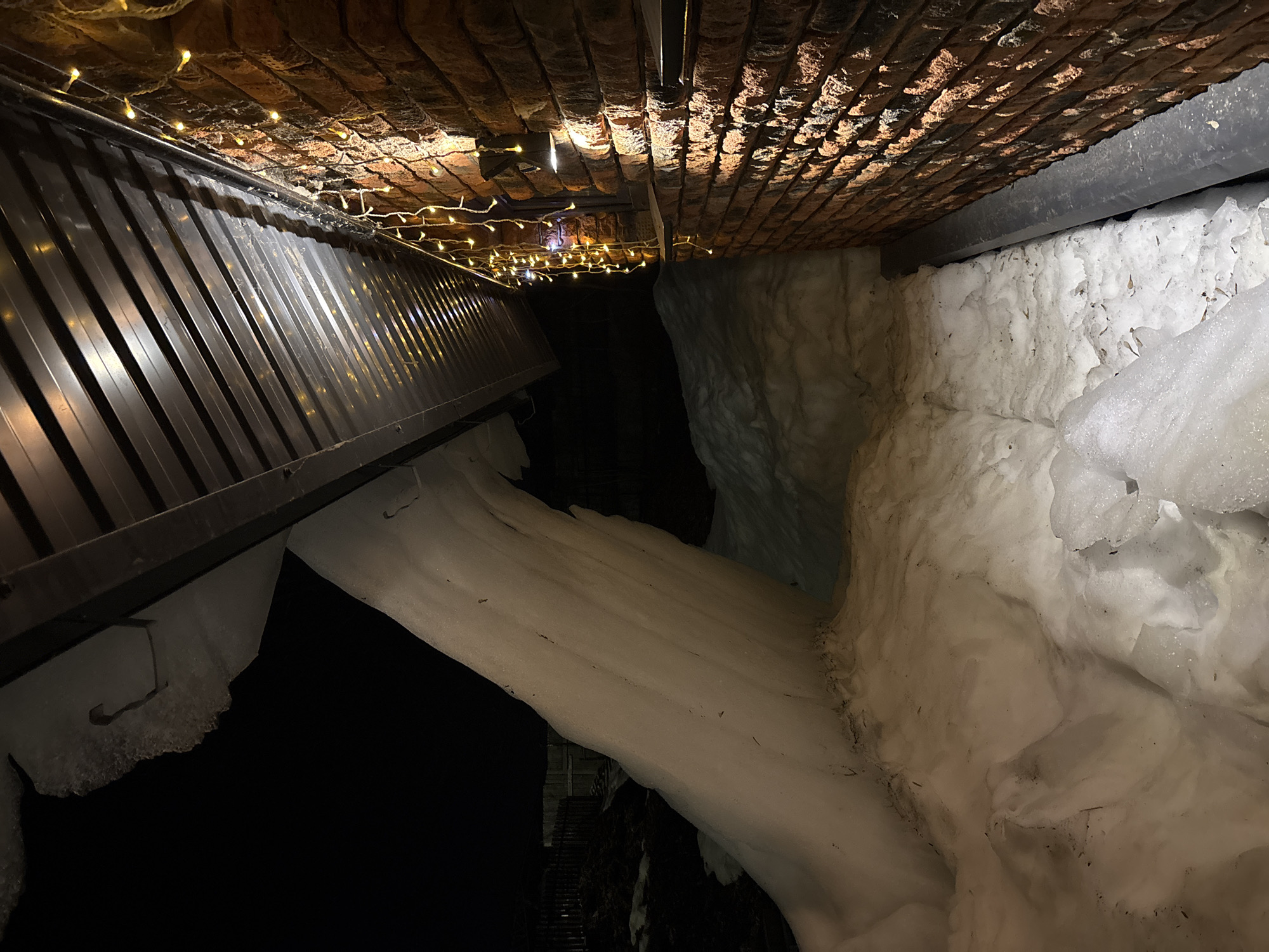 The image shows a constricted space under a brick ceiling adorned with fairy lights. To the left, a corrugated metal overhang with snow attached to its edge is visible. On the right, a large, irregular mass of snow fills the space, obscuring the view. The location is the city of Chaikovsky, Russia. The scene is dark, with most of the space beyond the snow-filled area appearing as a void, except for a hint of stairs in the distance.