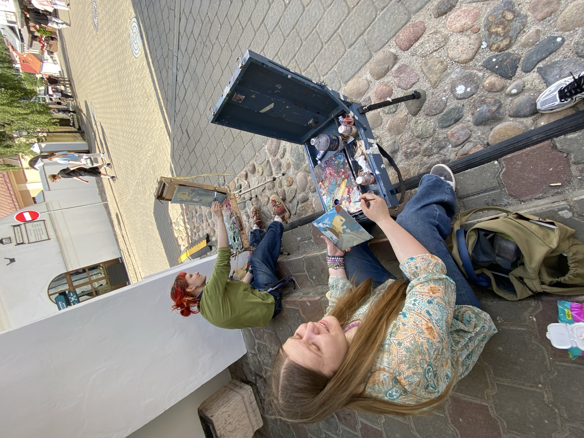 Two young women are painting outdoors on a cobblestone street. The woman on the left has bright red hair styled in a messy bun and is wearing a green long-sleeved shirt. She is leaning forward, focused on her canvas. The woman on the right has long brown hair and is wearing a patterned shirt. She is holding a smaller painting in her left hand and appears to be looking at her artwork.
In front of them is a portable art easel with paints and brushes. A large, open blue art box is on the ground, filled with tubes of paint and a palette. A green canvas bag is next to the art box. The scene is set on a sunny day, with shadows cast on the cobblestones, suggesting it is late morning or early afternoon. The architecture in the background, including white buildings and arched windows, hints at a European urban setting. The overall mood seems relaxed and creative, with the women engrossed in their artistic pursuits.