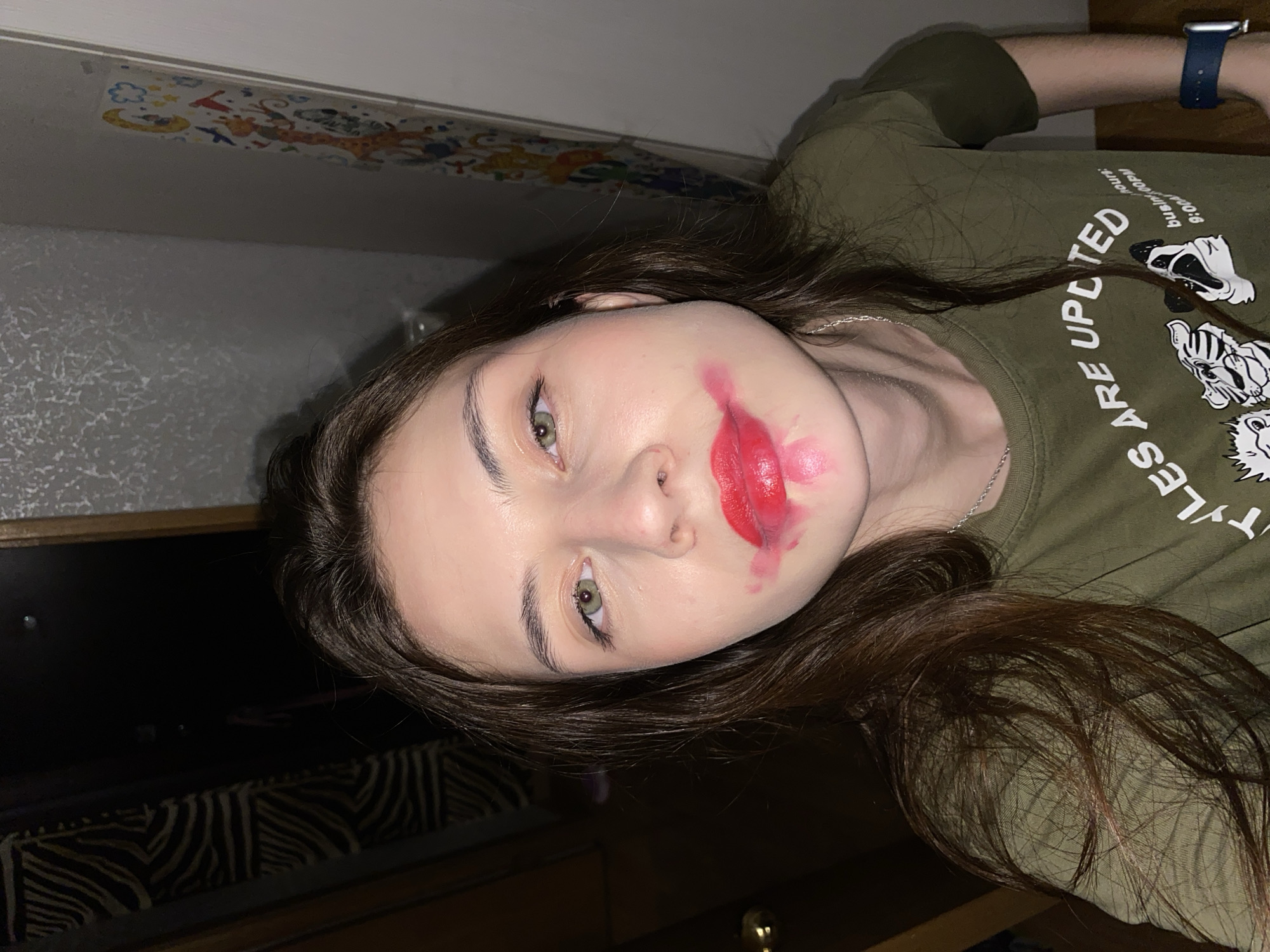 A photograph shows a young woman lying down indoors. She has fair skin, brown hair, and green eyes. Her lipstick is smeared around her mouth. She is wearing an olive green t-shirt with white writing and cartoon animal images. A silver chain necklace is visible around her neck. Her left wrist shows a blue smartwatch. The background shows a patterned wall and furniture. There is no information in the image to indicate that this scene is in Votkinsk, Russia.