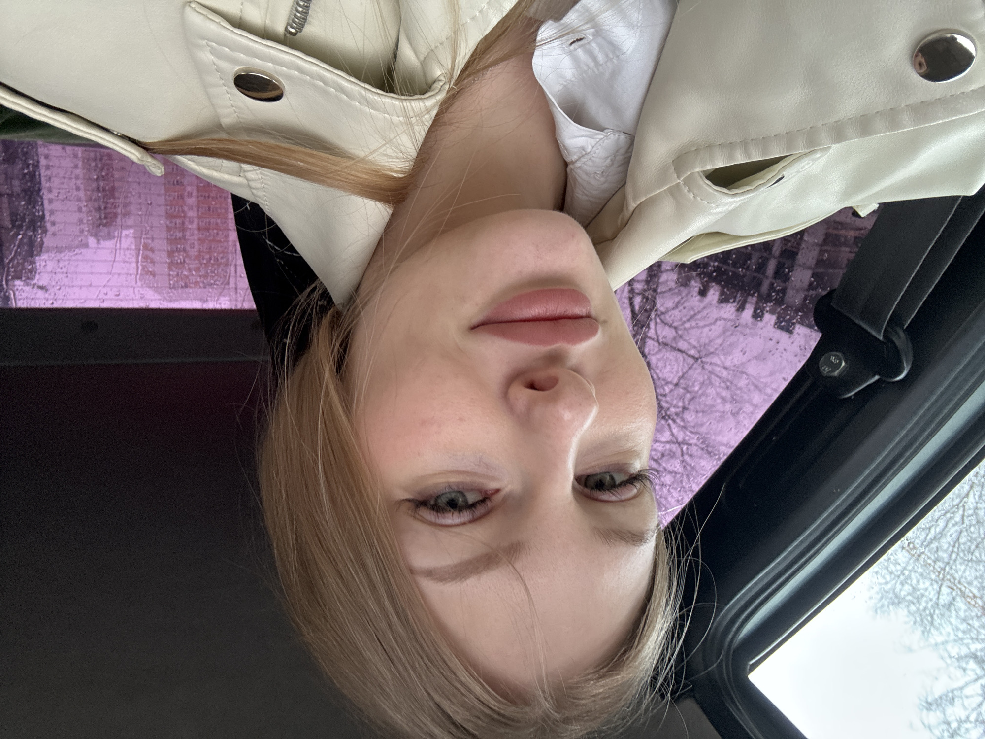 An upside-down portrait selfie features a young woman with long, light brown hair, lying or leaning back inside a car. She wears a cream-colored faux leather jacket over a white collared shirt. Her head and upper torso are visible, with her gaze directed neutrally towards the camera. Her lips are softly closed, and her green eyes are framed by subtle eyeliner and lashes.
The background reveals the interior of a car, including a black seatbelt mechanism on the right, which has a bolt with faint, unreadable text. Through the car windows, bare tree branches are visible, suggesting a cold season like winter or early spring. Rain or condensation is present on the window glass, further indicating wet weather. A distinctive purple tint covers a significant portion of the windows, possibly from a filter, unique window tinting, or a reflection, through which blurry outlines of buildings are seen, hinting at an urban or suburban environment. The overall lighting is soft and diffused, characteristic of an overcast day.