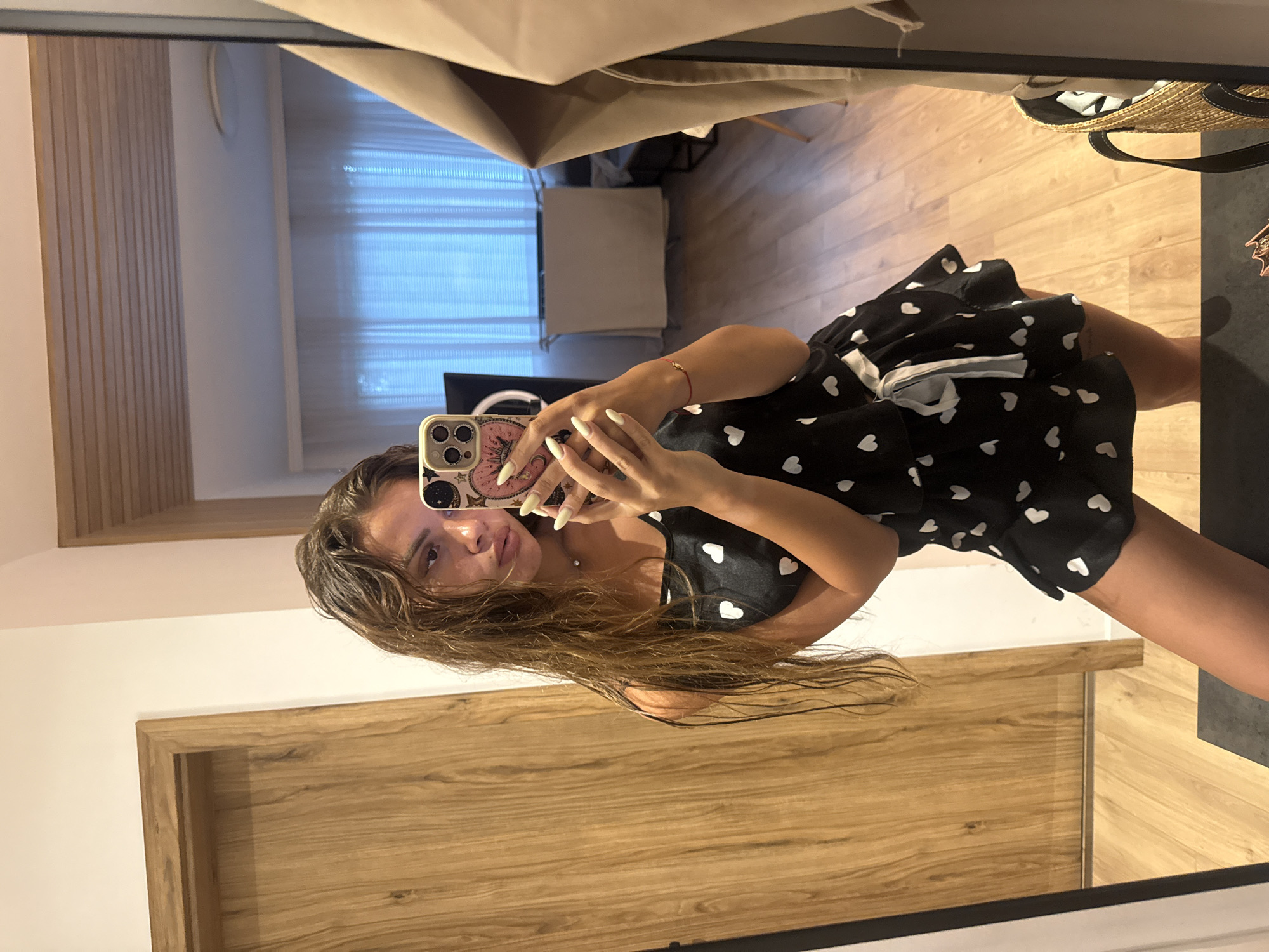 This is a media file captured in Plovdiv, Bulgaria. The image shows a young woman taking a selfie in a mirror. She has long, brown hair and is wearing a black dress with white heart patterns. She is holding a smartphone in her right hand, which has a colorful, decorated case. She is also wearing a thin red bracelet on her left wrist. The reflection in the mirror reveals a room with light-colored walls and wooden flooring. Behind her, vertical wooden panels are visible, as well as sheer white curtains covering a window. A straw bag with dark straps is placed on a dark surface to her right. The lighting suggests it is daytime.