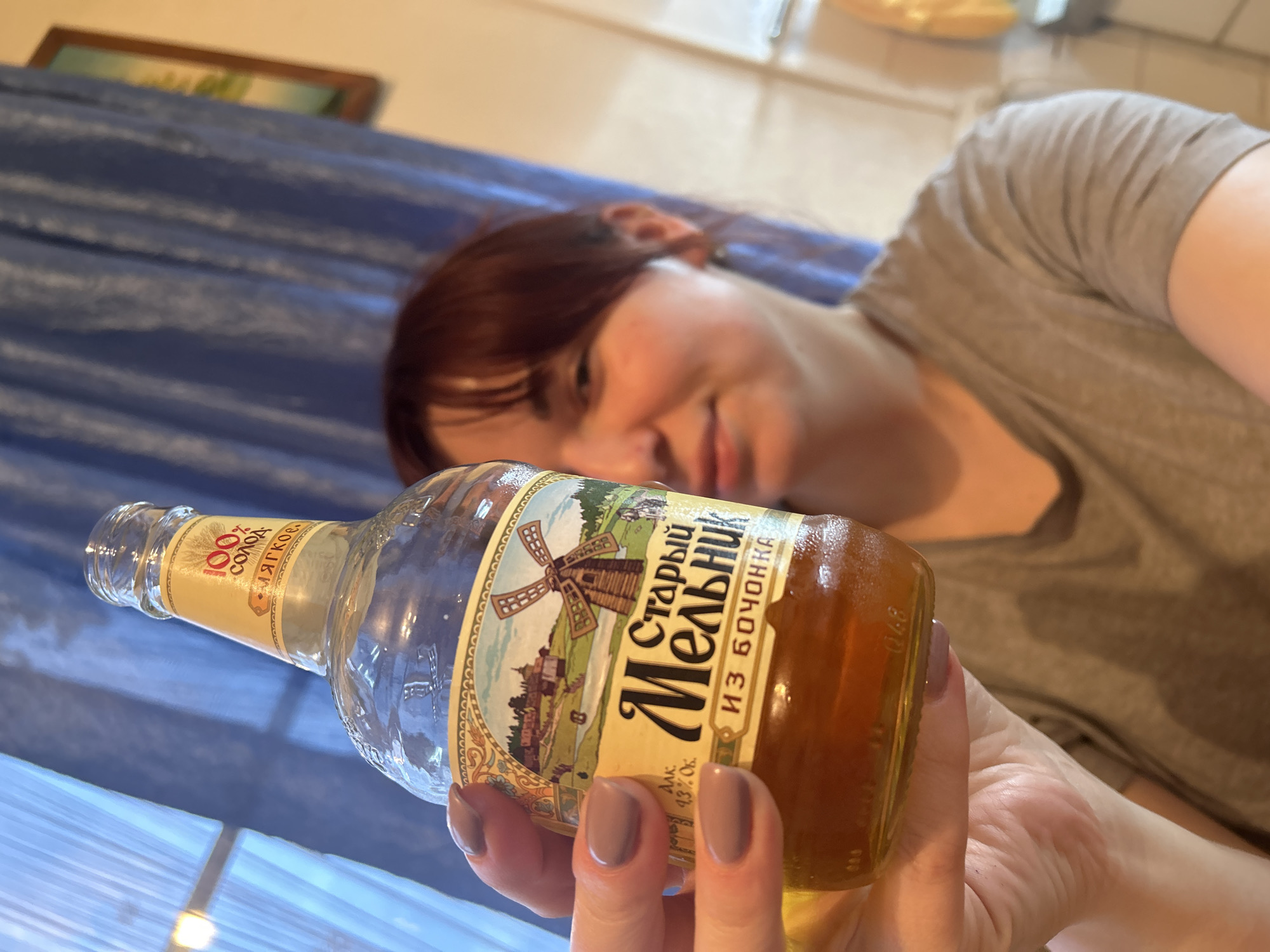 An indoor, close-up shot captured in Фоки, Russia, features a person holding a glass bottle with an amber liquid. The person, with reddish-brown hair and wearing a grey t-shirt, is mostly blurred in the background, appearing to be lying down or reclined, with a faint, relaxed expression. Their hand, with taupe-colored fingernail polish, holds the bottle prominently in the foreground.
The bottle's label is detailed, featuring a pastoral scene with a windmill. Visible text on the label includes "Старый Мельник" (Stary Melnik, meaning Old Miller) and "ИЗ БОЧОНКА" (IZ BOCHONKA, meaning FROM A BARREL), indicating it's likely a brand of alcohol, possibly a beer or spirit given the amber color and 43% alcohol content. Other text reads "100% СОЛОД" (100% MALT) and "Мягкое." (Myagkoye, meaning Smooth or Soft).
In the background, a blue curtain runs horizontally, and a blurry framed picture is visible above it. The lighting suggests it is daytime. The overall scene conveys a casual, relaxed atmosphere.