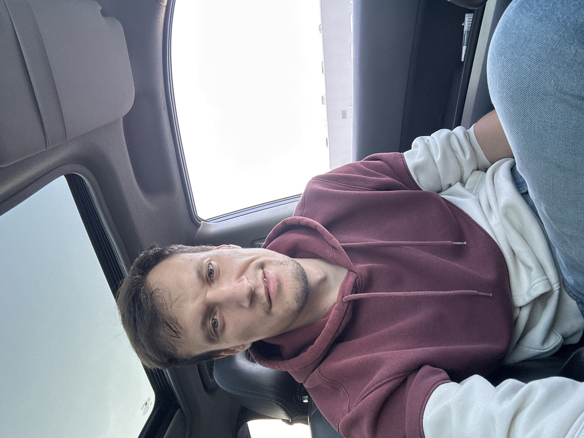 A man is seated in a car, looking directly at the camera. He has short, brown hair and light-colored eyes. He is wearing a maroon and cream-colored hoodie and blue jeans. The car's sunroof is open, revealing a bright, overcast sky. The interior of the car, including the headrest and dashboard, is visible. Outside the car window, a long, low building with a white facade can be seen, suggesting an industrial or commercial area. The scene appears to be in an outdoor setting, possibly a parking lot or roadside. There are no explicit visual cues to confirm the location as Kamianets-Podilskyi, Ukraine.