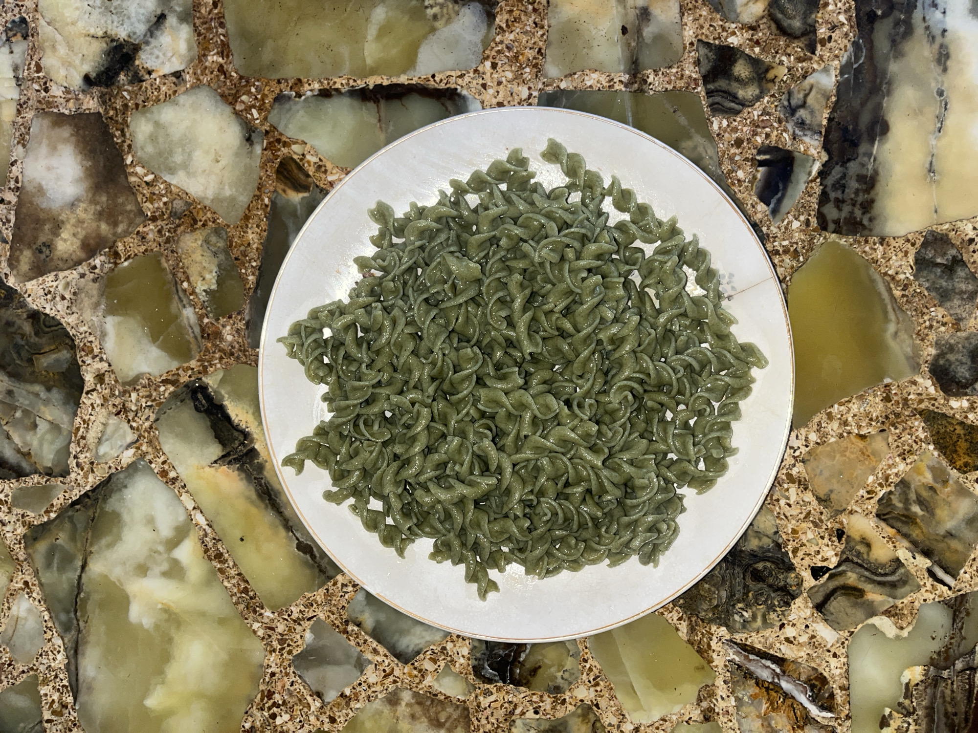 A top-down shot shows a white plate piled high with green pasta on a mottled, brown-and-tan stone surface. The pasta appears to be of the fusilli variety, though with a slightly different, less pronounced spiral. The exact type of stone surface is unknown, but it has a mosaic-like pattern and would appear to be inside a residential location. It is impossible to confirm if the location is in Vicente López, Argentina, based on the visual information provided. There are no people in the image, and the primary subject is the pasta. The action is the presentation of food on a plate. No interactions are visible.