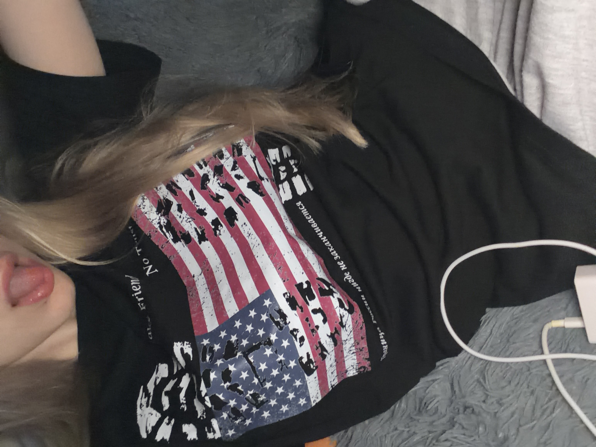 A person with blonde hair is visible from the chest up, wearing a black t-shirt with a distressed American flag graphic. The person's tongue is sticking out. The t-shirt also has text, which appears to be in Russian, below the flag. A white charging cable and adapter are visible in the lower left of the image, lying on a grey fuzzy surface. The background is dark and appears to be a bed or couch. The image is an indoor shot, likely taken in a residential setting.