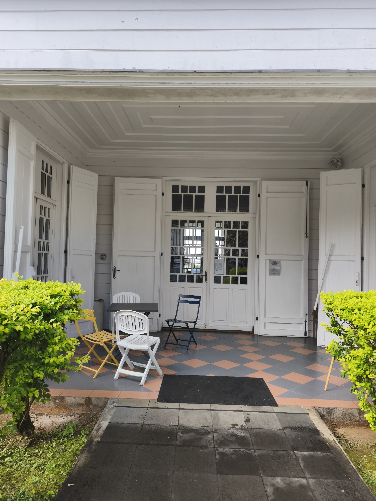 The image shows the entrance to a white wooden building with large double doors and shutters. A checkered tile floor with orange and grey squares is visible in front of the entrance, along with a black mat. Several chairs are placed on the tiled area, including two white plastic chairs, a yellow metal chair, and a blue metal chair. Bright green bushes flank the entrance on both sides. The building appears to be located in a residential area, possibly a home or a small business, in La Plaine-des-Palmistes, France. The architecture suggests a colonial or tropical style. The scene is static, with no people or animals present. The lighting indicates it is daytime.