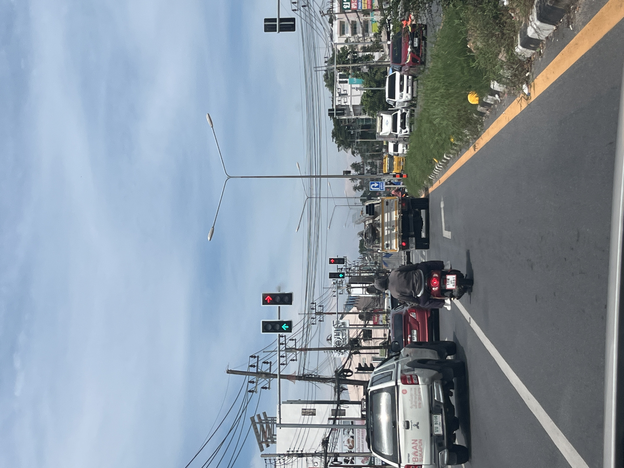 The image depicts a traffic scene on a multi-lane road in Rawai, Thailand, under a partly cloudy sky. In the foreground, a white pickup truck is visible from the rear left, displaying a license plate that reads "UU 8006" and "BAAN SUKAPUN". To its right, a red car with license plate "VS 3749" is positioned slightly ahead. A person is riding a red scooter with a passenger behind them, their license plate reads "108 5031".
Further down the road, traffic lights are visible. The closest set shows a green arrow pointing left and a red arrow pointing right, indicating traffic signal status. Further ahead, another traffic signal shows a red light. Several vehicles are stopped or moving slowly in traffic, including a yellow truck, white trucks, and white SUVs.
The environment includes power lines crisscrossing overhead, supported by numerous utility poles. Buildings line the sides of the road, some appearing to be commercial establishments with signage in Thai script. Green vegetation and grass grow along the roadside, with concrete barriers and a yellow line marking the edge of the road. The overall impression is a typical busy intersection in a tropical urban environment.