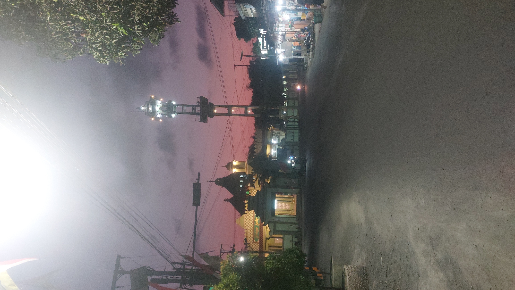 The image captures a street scene in Sedan, Indonesia, during twilight. In the center of the frame, a tall, illuminated tower stands prominently, with lights adorning its structure. To the left, a mosque complex is visible, featuring distinctive architecture with multiple domes and an entrance gate. The mosque's facade has Arabic inscriptions. The sky displays a gradient of purple and pink hues, indicative of sunset or dawn. A paved road occupies the foreground, with a dirt shoulder on the left side where planters are situated. Several streetlights illuminate the scene, and utility poles with wires are present. On the right side of the road, shops with awnings and lights are visible. A few motorcycles and a person on a motorcycle are present on the street, suggesting ongoing activity. A tree with dense foliage stands on the far right. The overall atmosphere is serene, with the artificial lights creating a contrast against the fading natural light.