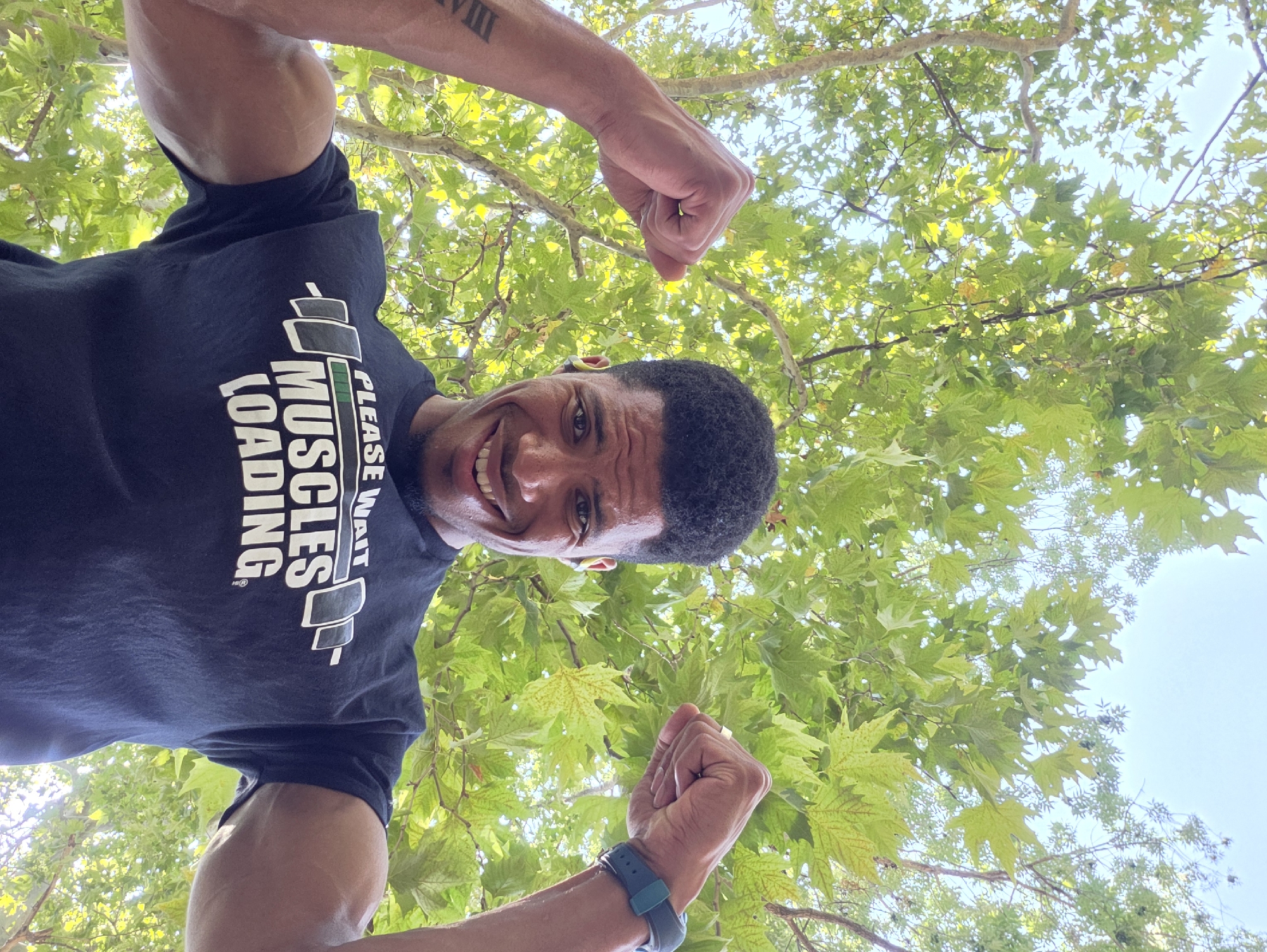 A man is posing for a selfie in front of a tree. He is smiling and flexing his biceps. He is wearing a black t-shirt with the words "Please Wait Muscles Loading" printed on it. The man has dark curly hair and is wearing a fitness tracker on his left wrist. The background is a canopy of green leaves against a bright sky. The scene appears to be outdoors in a park or a garden. The man's right arm is blurred due to motion. The man's left arm has a tattoo of Roman numerals visible. There is no information about the location in Varna, Bulgaria.