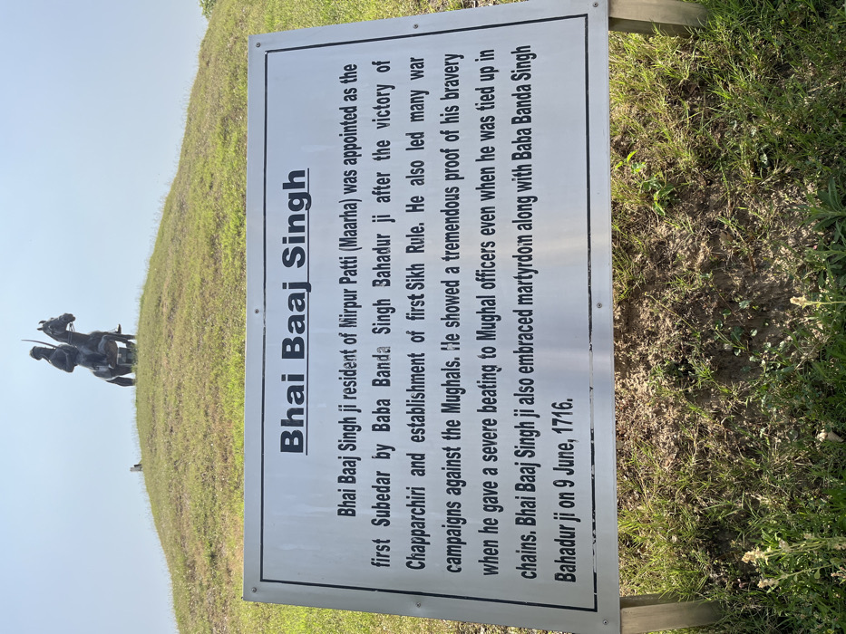 A metal sign with the words "Bhai Baaj Singh" in large bold font stands in front of a grassy mound. The sign contains text about Bhai Baaj Singh, a resident of Mirpur Patti (Maarha), who was appointed as the first Subedar by Baba Banda Singh Bahadur ji after the victory of Chapparchiri and establishment of first Sikh Rule. He also led many war campaigns against the Mughals. He showed a tremendous proof of his bravery when he gave a severe beating to Mughal officers even when he was tied up in chains. Bhai Baaj Singh ji also embraced martyrdom along with Baba Banda Singh Bahadur ji on 9 June, 1716. The sign is situated on a grassy mound, the top of which is capped with a statue of a man on horseback. The statue faces directly forward. The scene is taken in Sahibzada Ajit Singh Nagar, India (latitude: 30.703, longitude: 76.668). The sky is bright blue and clear with no clouds. There are no people visible in the image.