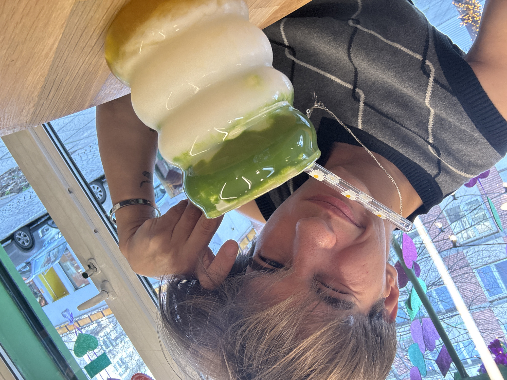 A male, likely in his late teens or early twenties, wearing a gray and black argyle vest and silver necklace, holds a layered drink with a straw in front of his face. The drink is layered with yellow on top, white in the middle, and green on the bottom. He is positioned next to a window with views of a street scene. Through the window, parked cars, buildings with colorful painted facades, and decorations that resemble leaves or flowers are visible. The location appears to be in an urban setting, possibly in Sofia, Bulgaria, based on the visible architecture and street decorations.