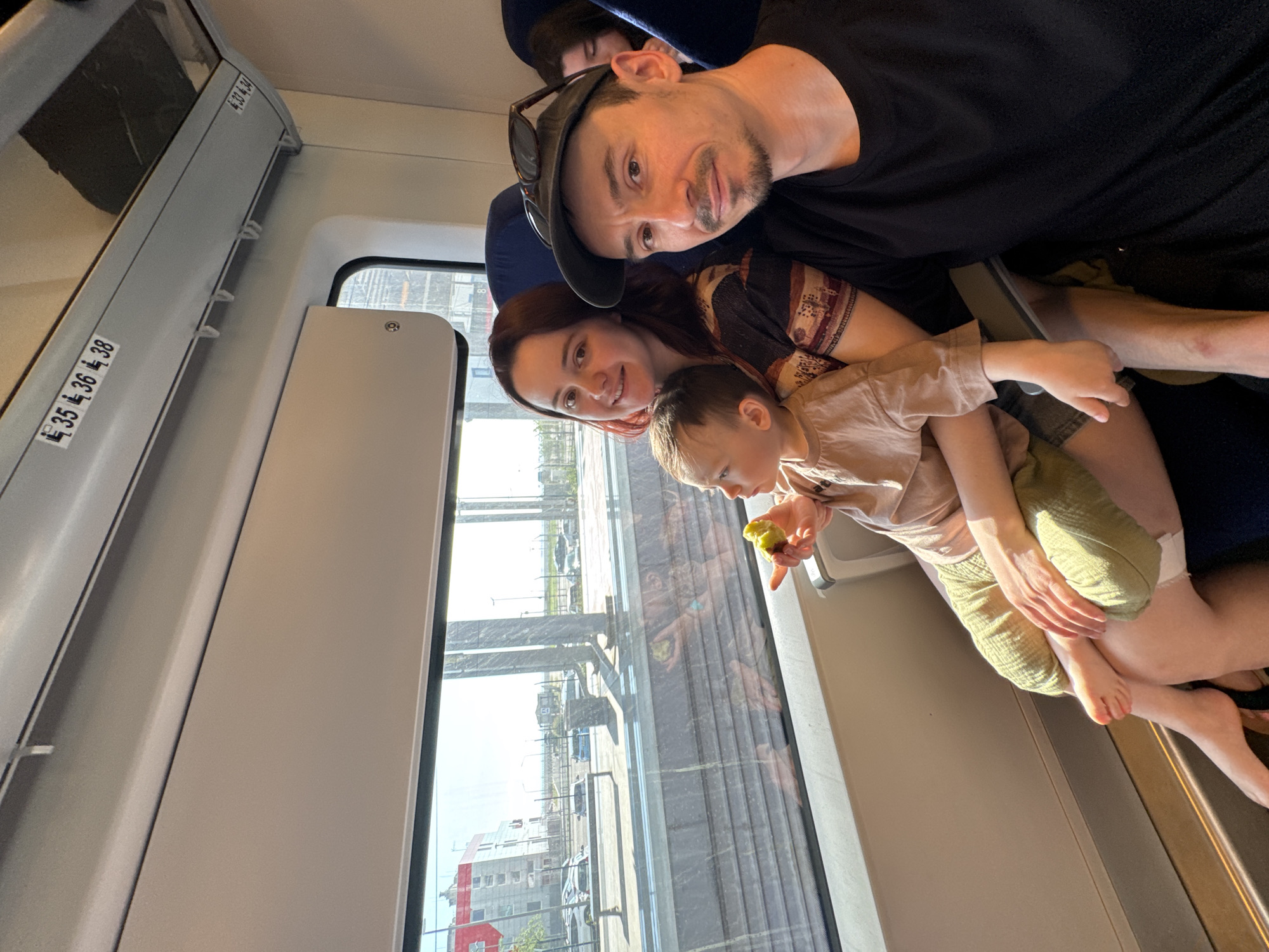 The image shows a family of three on a train, likely traveling within the city. The train car is modern, with large windows offering a view of the cityscape. Outside the window, buildings and infrastructure are visible, characteristic of a railway approach to a city. The family appears to be taking a selfie. The father is wearing a cap with sunglasses on top, a black shirt, and shorts, while the mother is in a patterned top. Their child is holding what appears to be a piece of fruit, possibly an avocado, and is wearing a light-colored shirt and shorts. The train car's interior is bright and modern, with seat numbering visible above. The family is the main subject of the image, capturing a moment during their train journey. Based on the style of the architecture and vegetation visible through the window, the location is consistent with Sochi, Russia.