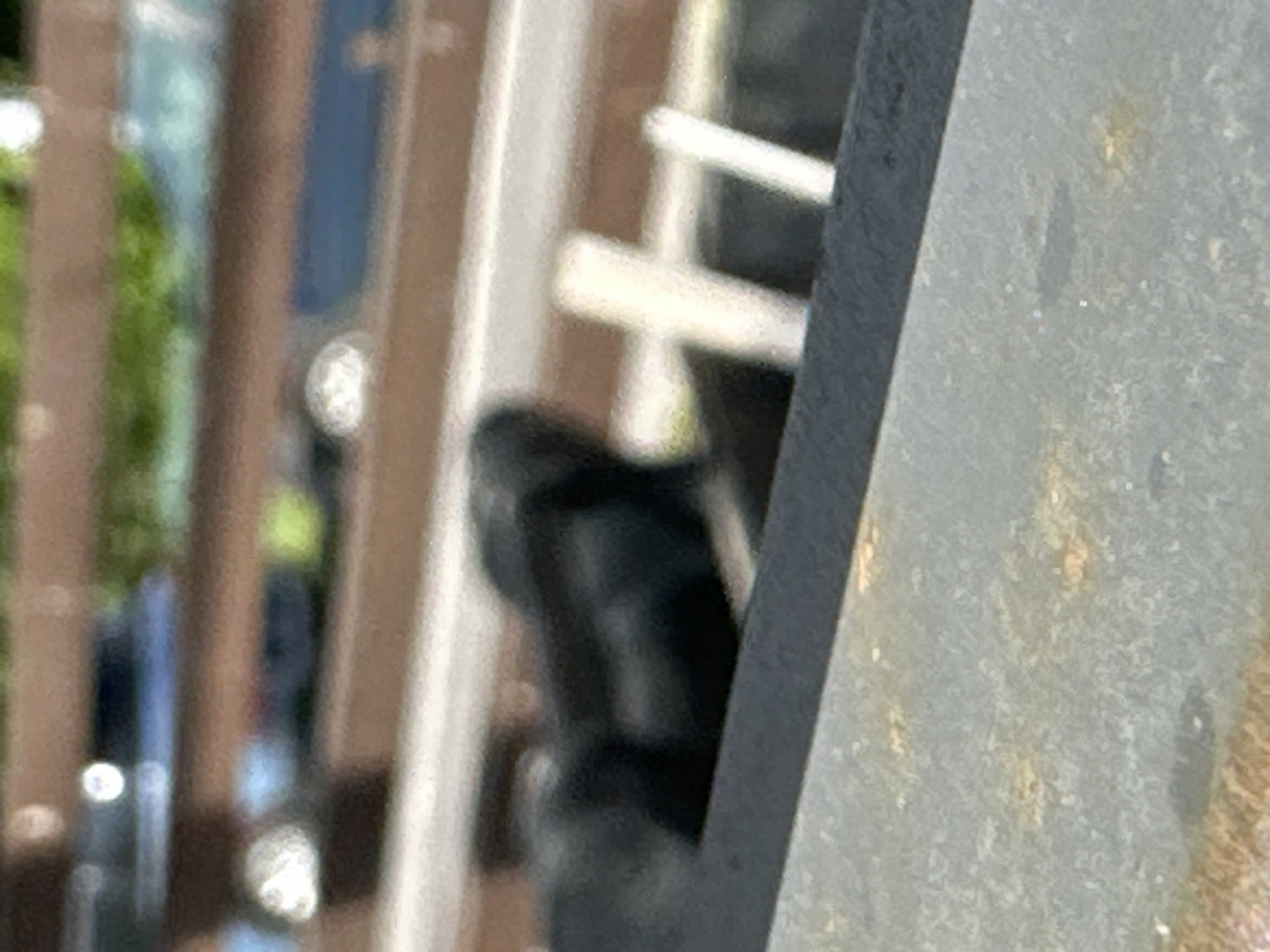The image is a close-up, out-of-focus shot of a black object that appears to be a car tire or a similar rubber item. It is positioned within a dark rectangular opening or container. In the background, there are blurred shapes of vehicles and some greenery, suggesting an outdoor urban setting. The foreground shows a rough, textured surface that is likely metal, possibly part of a railing or a bench. The overall impression is of an industrial or public space with vehicles present. There are no discernible features to determine a specific location within Minsk, Belarus.