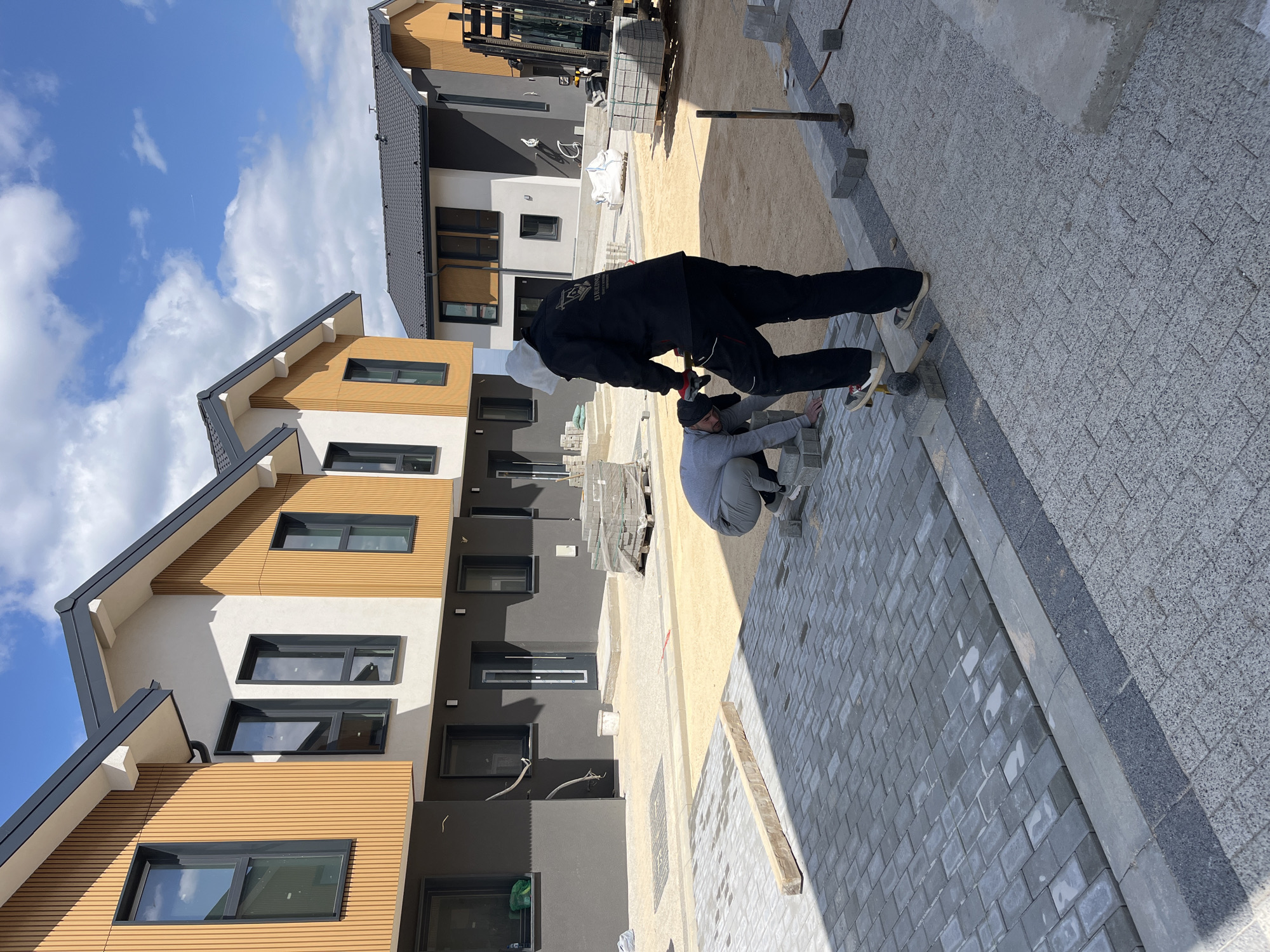 The image shows two individuals laying pavement stones in Branipole, Bulgaria. They are working on a street lined with newly constructed, multi-story residential buildings. The buildings have a modern design with a combination of wooden cladding, white and dark grey facades, and black window frames. There is a forklift parked near one of the buildings, and stacks of paving stones are visible. The sky is blue with scattered clouds.