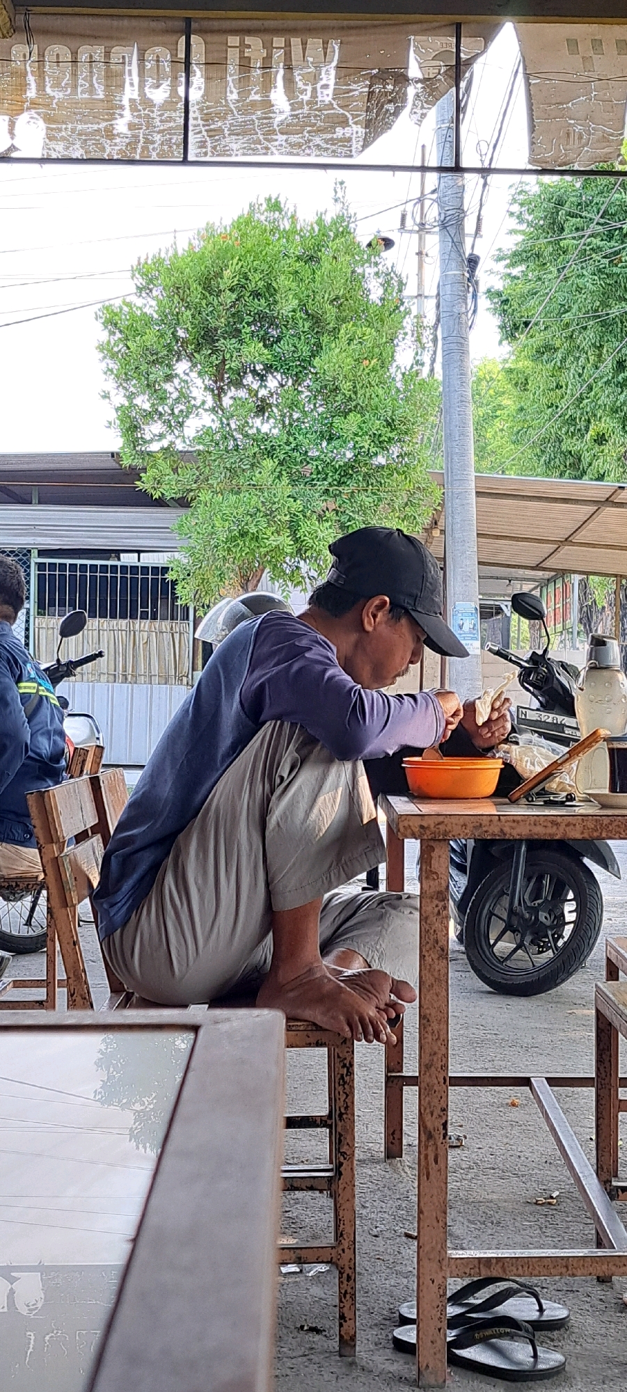 A man wearing a blue long-sleeved shirt, khaki pants, and a black baseball cap is seated on a wooden chair outside in Nogosari, Indonesia. He is hunched over a table, holding a piece of food in his right hand and a utensil in his left, appearing to eat from an orange bowl. A motorbike is parked behind the table, with a woman in a blue and yellow jacket visible in the background to the left. The environment appears to be an outdoor seating area with a corrugated metal roof overhead and trees and utility poles visible in the background. Flip-flops are on the ground under the table.