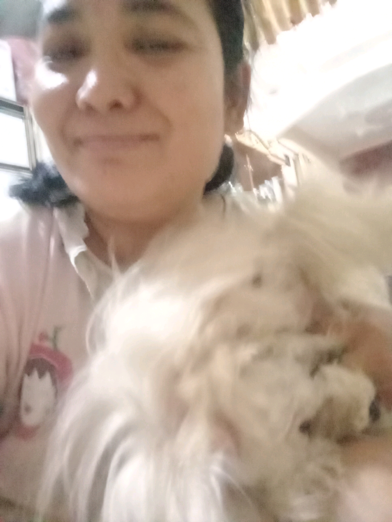 The image is a selfie taken indoors, likely in a residential setting in Jambi, Indonesia. A young woman is holding a small, fluffy white dog close to her face. The woman is smiling and looking towards the camera, while the dog is mostly obscuring her face and chest. The background is blurred and appears to be an interior space with some lighting and furniture visible, suggesting a casual, everyday scene. The lighting is bright, possibly from natural light sources or indoor lamps. The image quality is poor, with a noticeable blur and low resolution, making it difficult to discern fine details of the environment or the subjects.