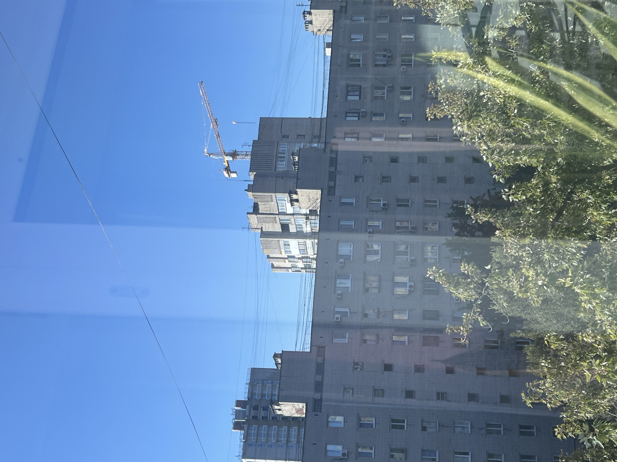 The image depicts a cityscape viewed through a reflective surface, likely a window. The dominant feature is a multi-story apartment building constructed from grey brick, characterized by numerous windows and visible air conditioning units. Behind this building, another, taller apartment building is under construction, showing exposed concrete and scaffolding. In the distance, a construction crane stands against a clear blue sky. Power lines are strung across the scene, intersecting the buildings. In the foreground, lush green trees partially obscure the view of the apartment building. The overall impression is one of urban development and residential areas in Volgograd, Russia.
