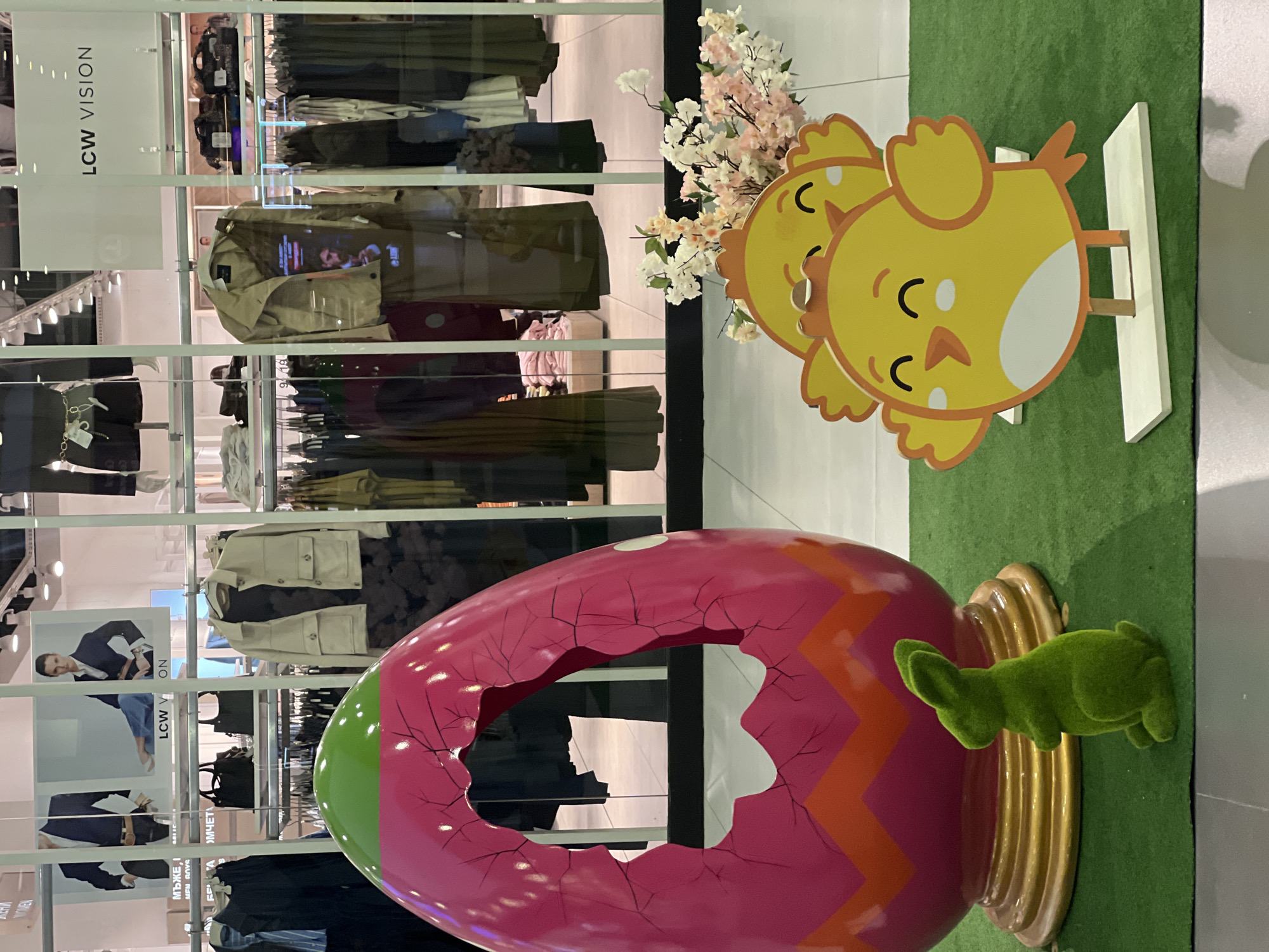 A close-up shot indoors displays Easter decorations in front of a storefront window with clothing on display at LCW Vision, likely located in Plovdiv, Bulgaria. An oversized, pink, cracked Easter egg with a green top and orange zigzag pattern is positioned on a gold-colored base with a green, moss-like bunny resting against it. Behind and above the egg, there is a standee of two yellow chicks with closed eyes and white bellies against a background of green faux grass. Behind the decorations, the store's window displays clothing on hangers and posters of models in suits and casual wear, visible signs with the name "LCW Vision," and Bulgarian text "МЪЖЕ, І MEN ROVS ТА ОМЧЕТА". A staircase and more clothing racks can be seen within the store, and light shines through the window, suggesting a bright, retail environment.