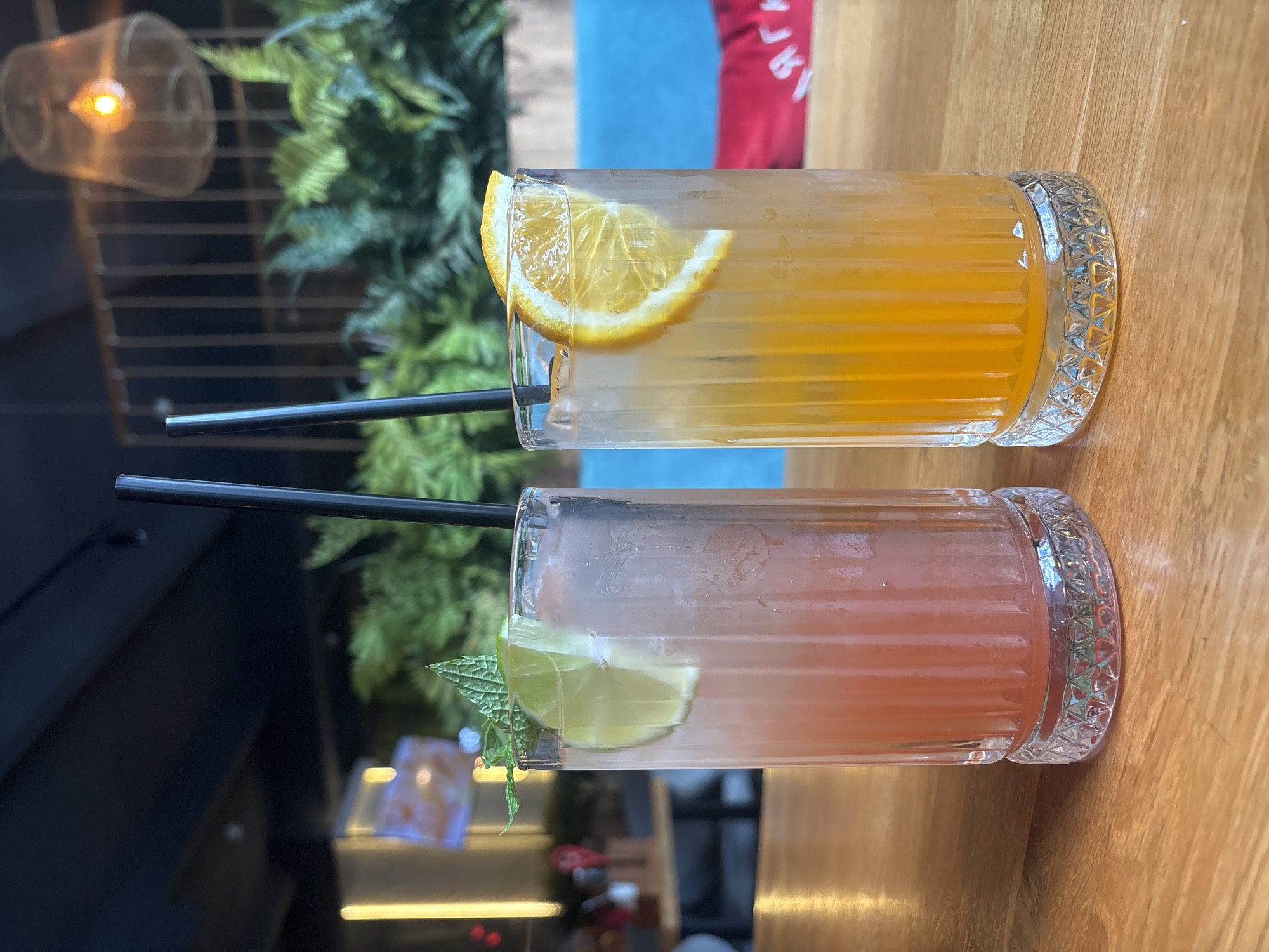 Two tall, fluted glasses filled with colorful cocktails sit on a wooden table. The glass on the left contains a pinkish-red beverage, garnished with a slice of lime and a sprig of mint. The glass on the right holds a vibrant orange drink, decorated with a slice of orange. Both cocktails have black straws. The background is softly blurred, showing hints of indoor foliage and a lamp fixture. There is no identifiable information about the city of Ivanovo, Russia, present in the image.